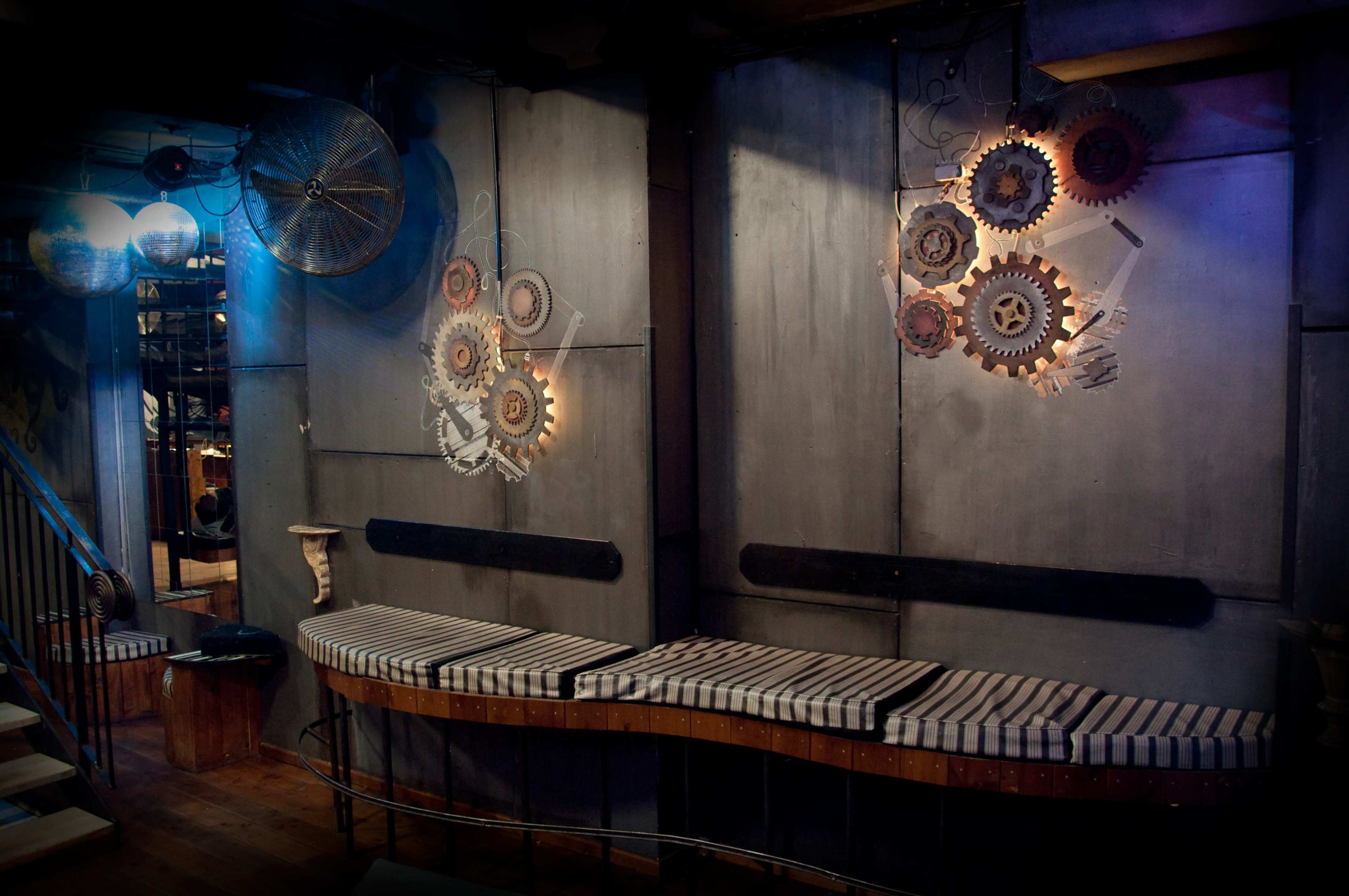 The image shows a dimly lit interior space featuring metal walls adorned with artistic gear designs and a striped seating area along one side.