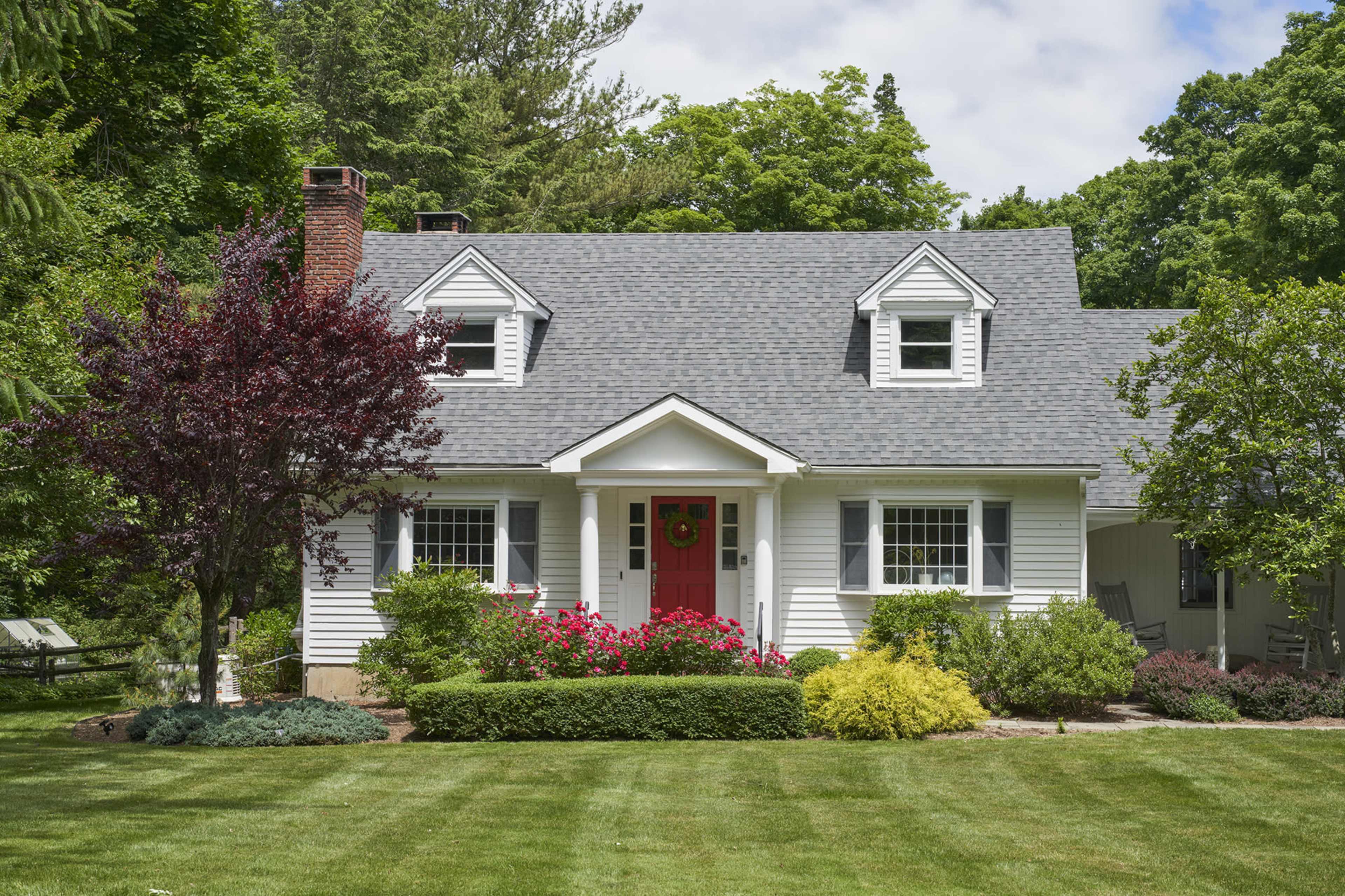 A two-story white house with a red front door is surrounded by landscaped greenery and flowering bushes.
