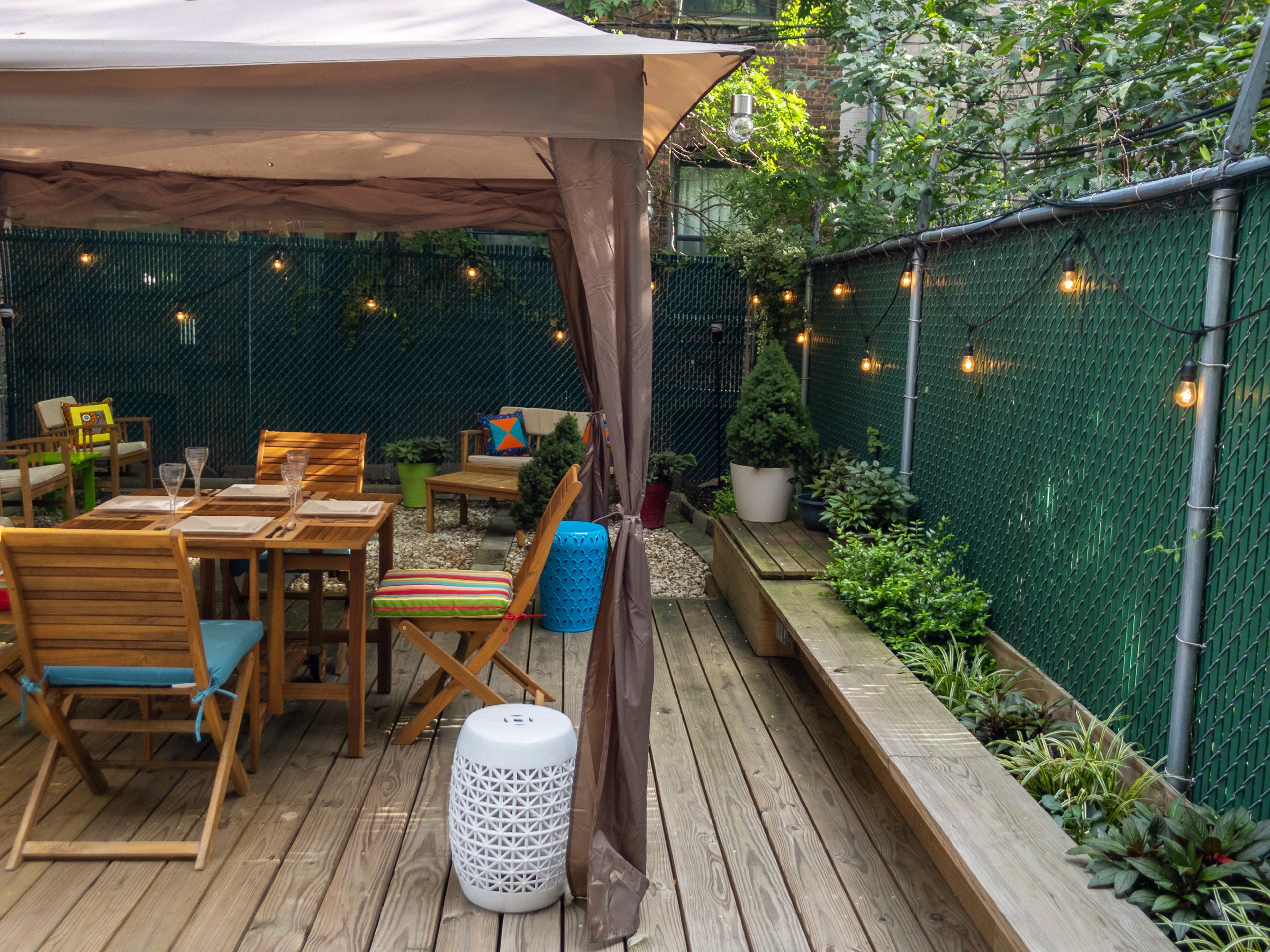 A wooden deck area with a table and chairs under a canopy, surrounded by greenery and string lights against a chain-link fence.