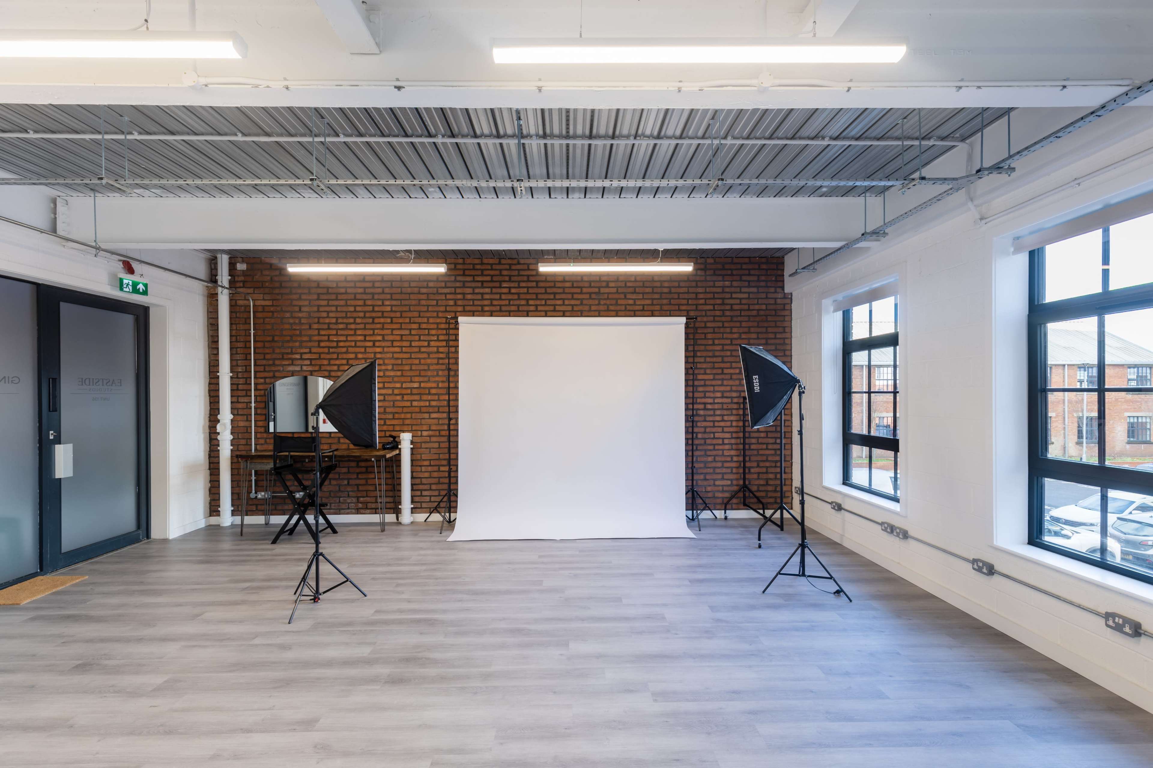 The image shows a photography studio with a blank white backdrop, two light stands, and large windows.