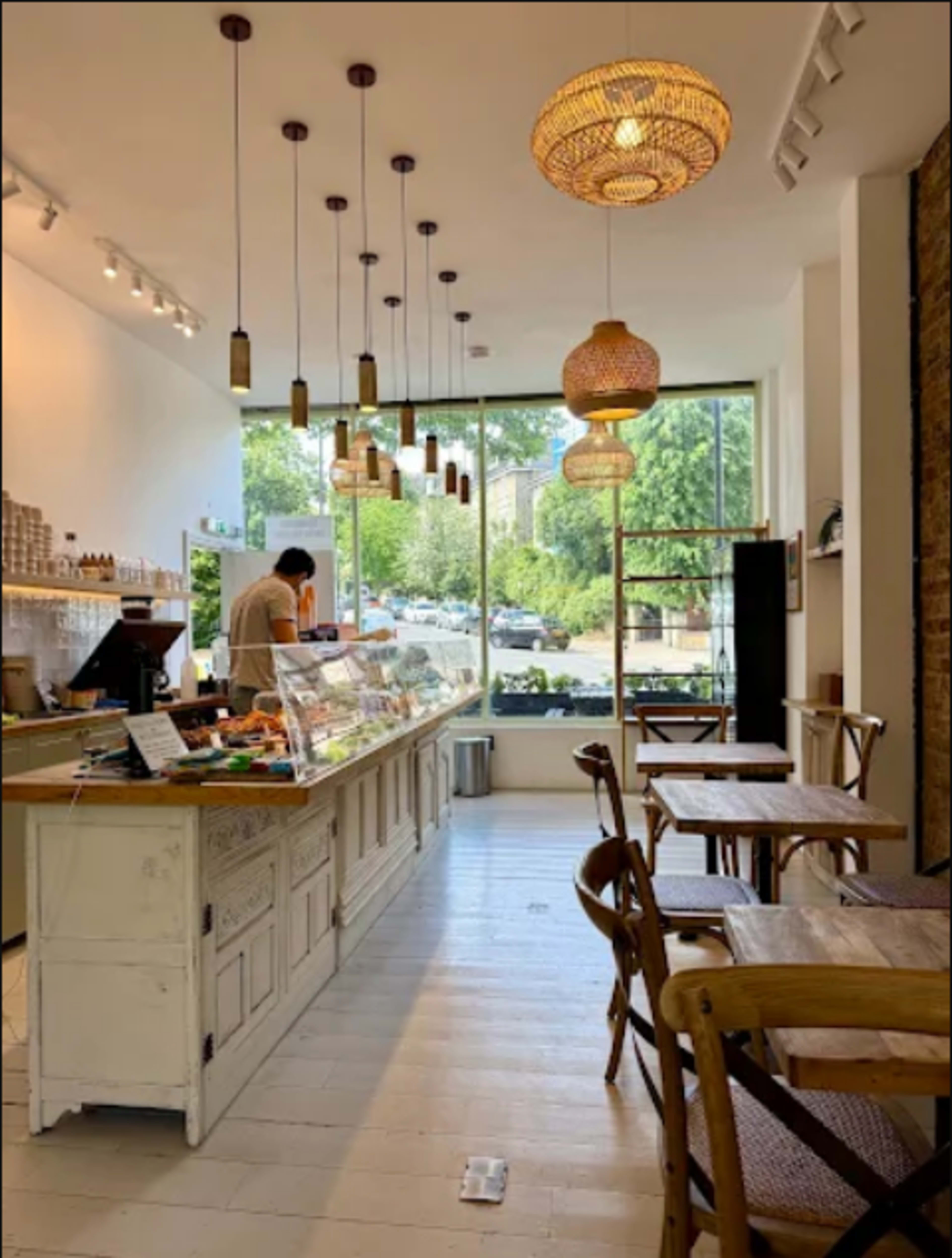A light-filled café features a wooden counter displaying pastries, with several tables and pendant lights hanging overhead.