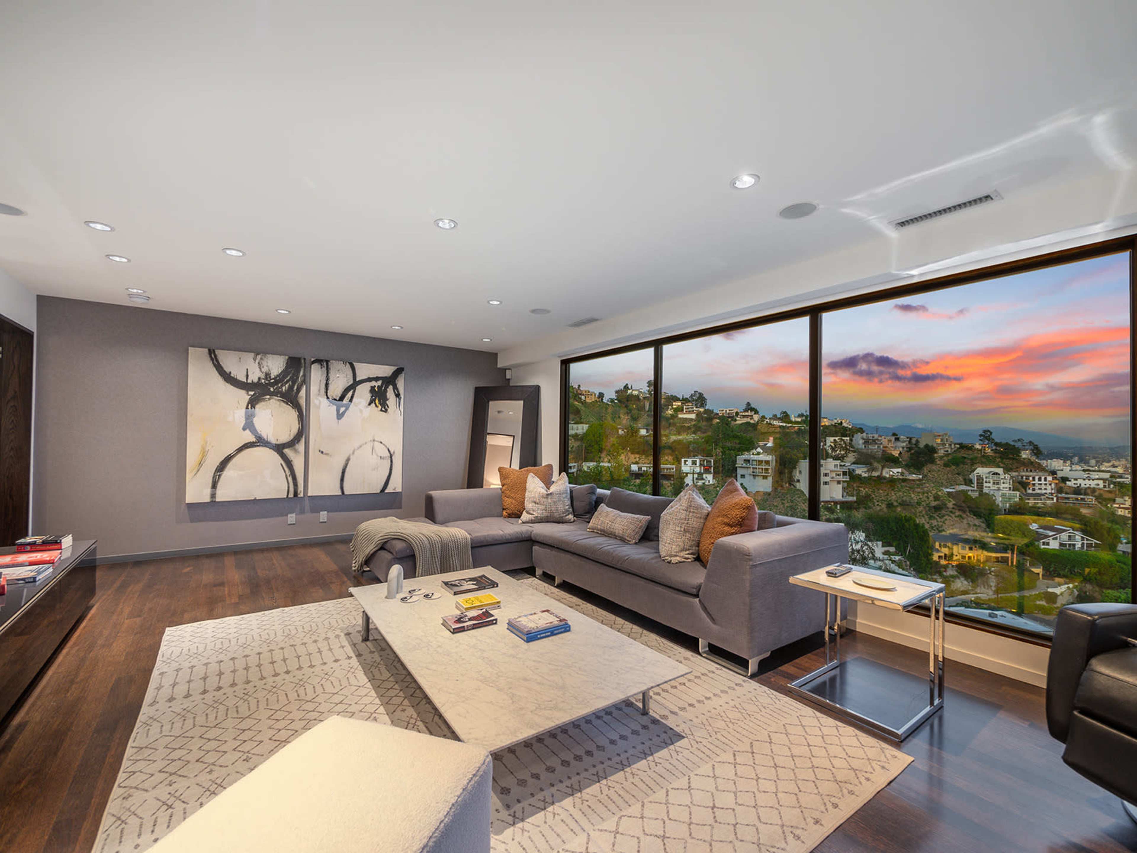 A spacious living room features a large sectional sofa, a coffee table, and expansive windows offering a view of a hillside at sunset.