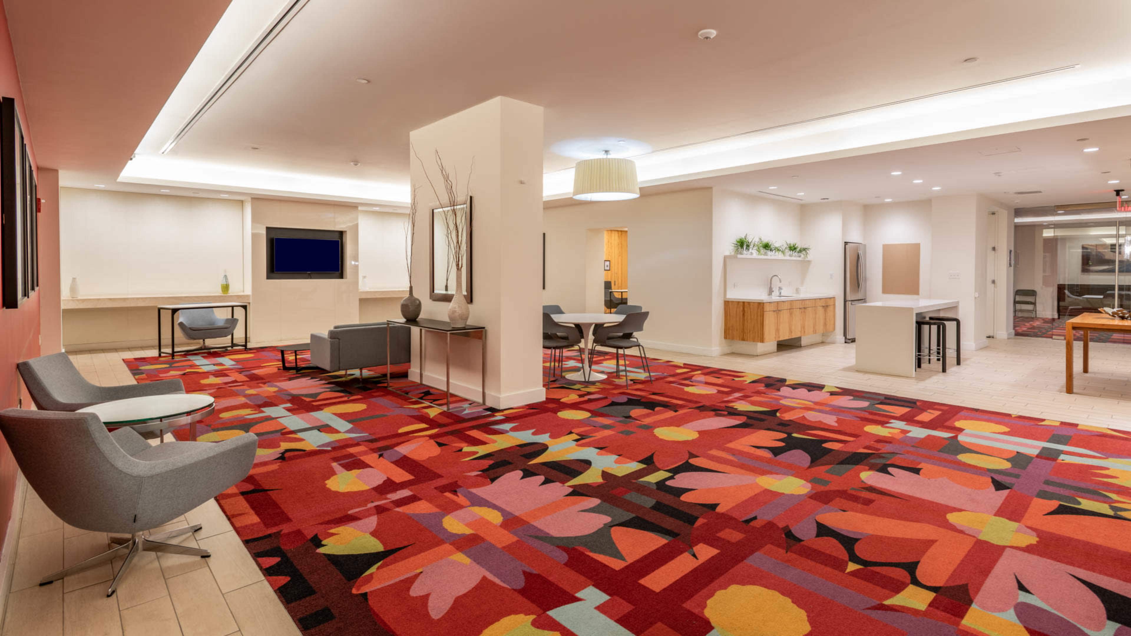 The image shows a modern lobby area with colorful floral carpet, seating arrangements, a television, and a kitchenette in an upscale interior space.