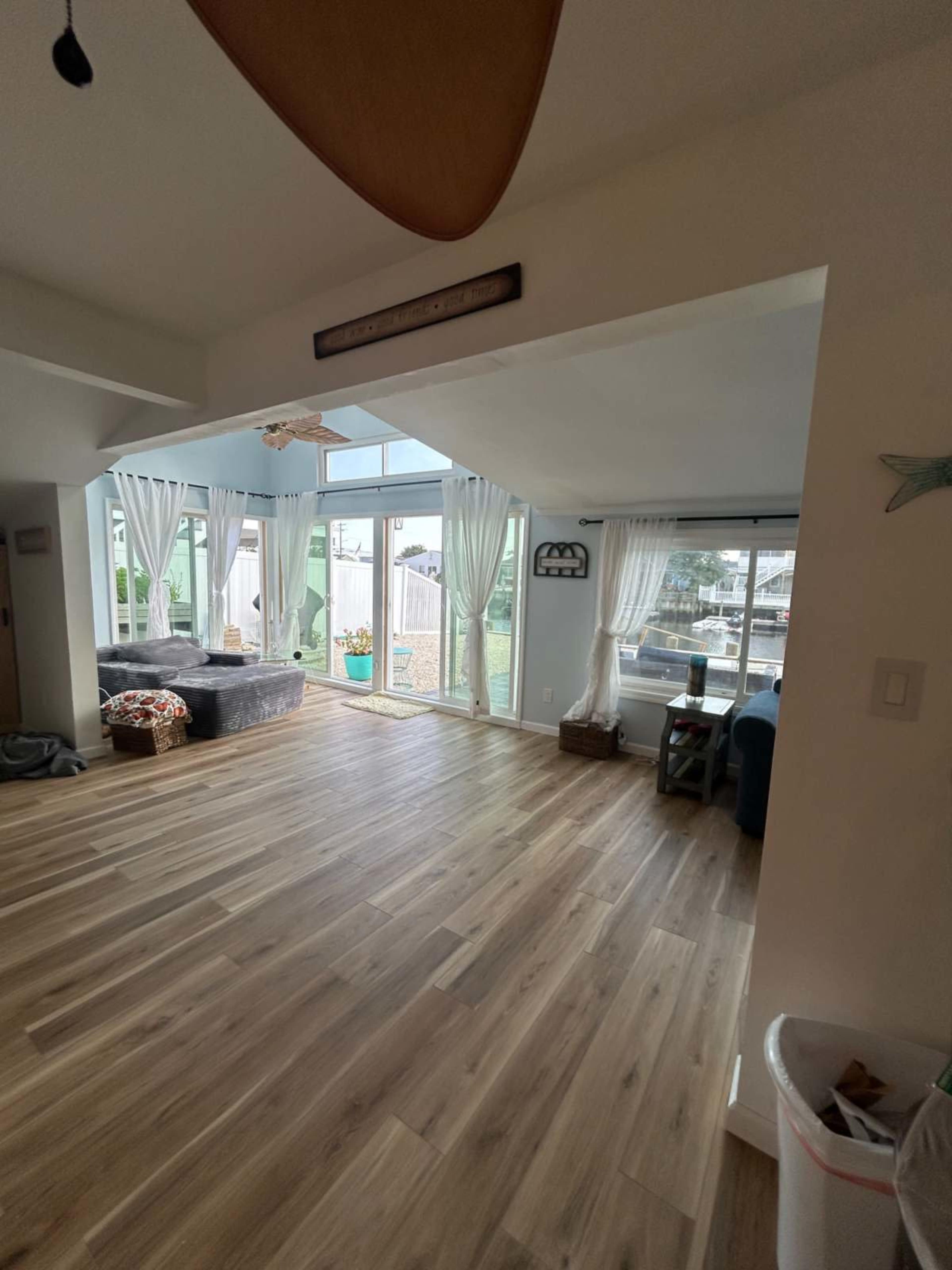 The image shows a brightly lit living room with wooden flooring, large windows, and light-colored curtains.