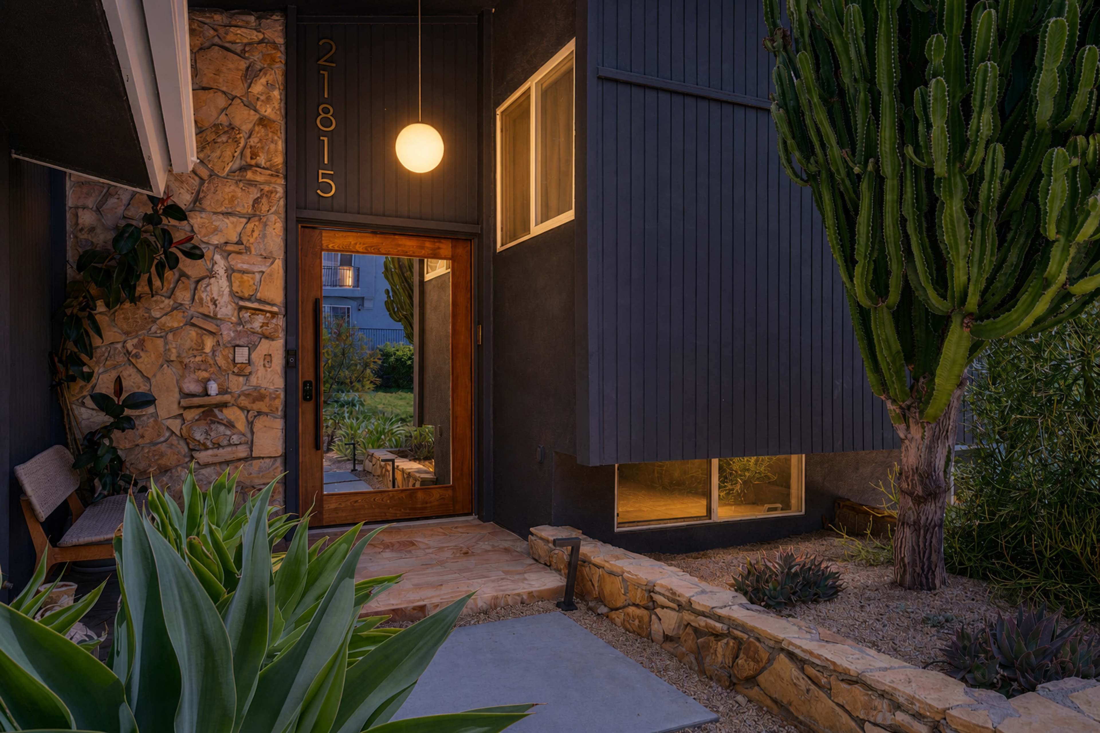 The image shows the entrance of a modern home featuring a wooden door, stone accents, and a large cactus beside the pathway.