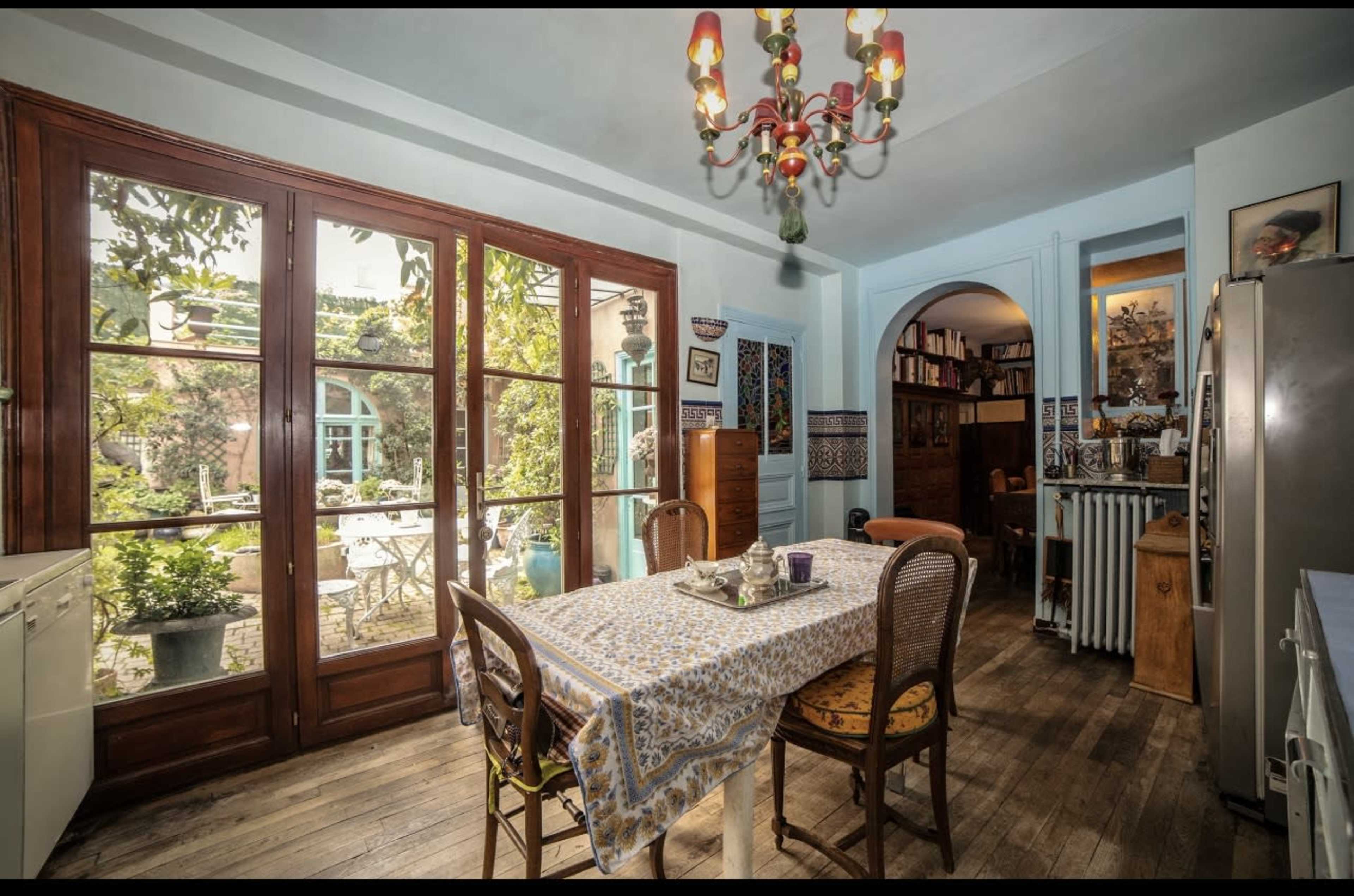 A spacious kitchen features a dining table set with a tablecloth, surrounded by chairs, with large windows showcasing a garden outside.