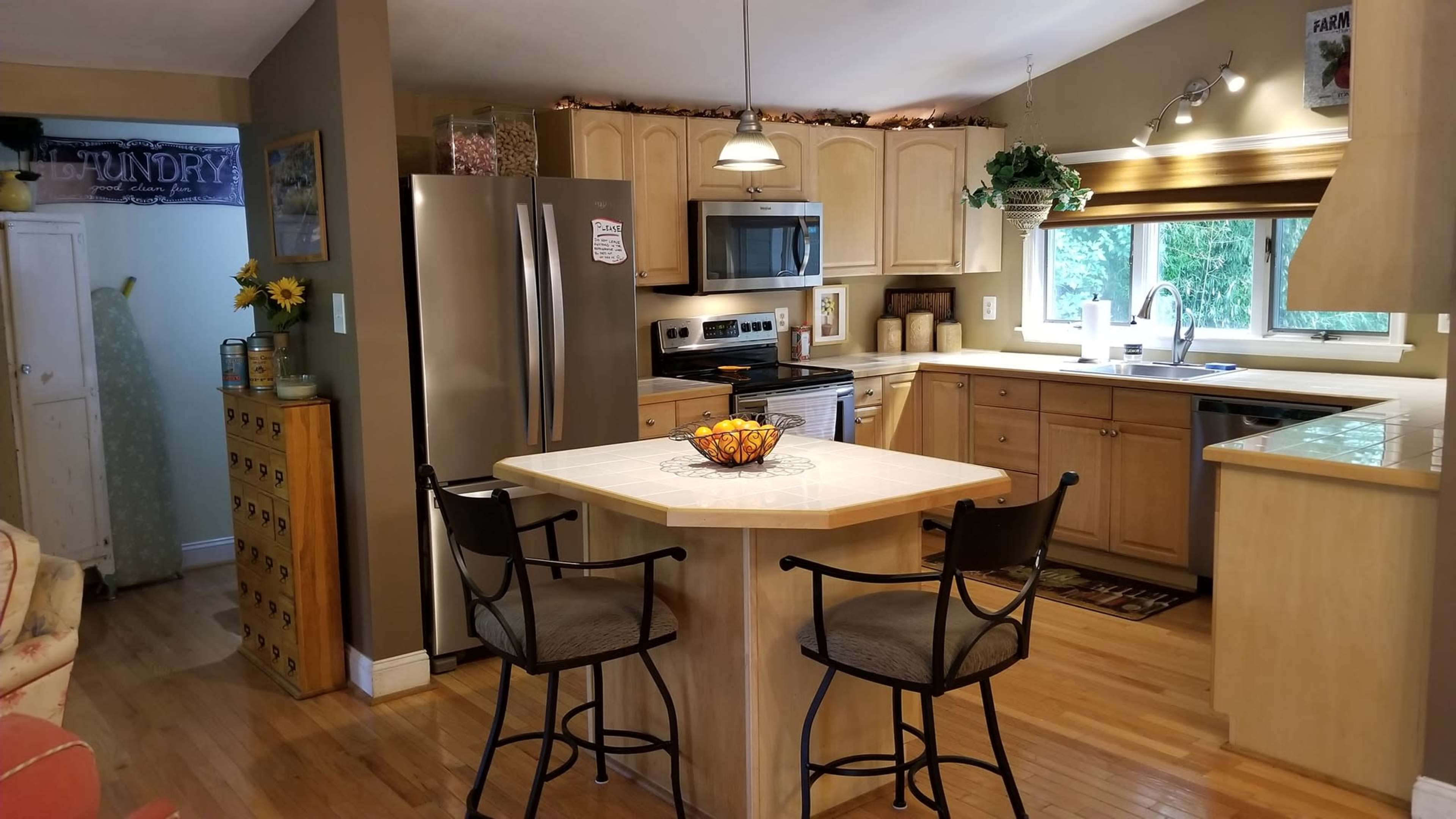 The image shows a well-lit kitchen with wooden cabinetry, stainless steel appliances, and a central island with bar stools.