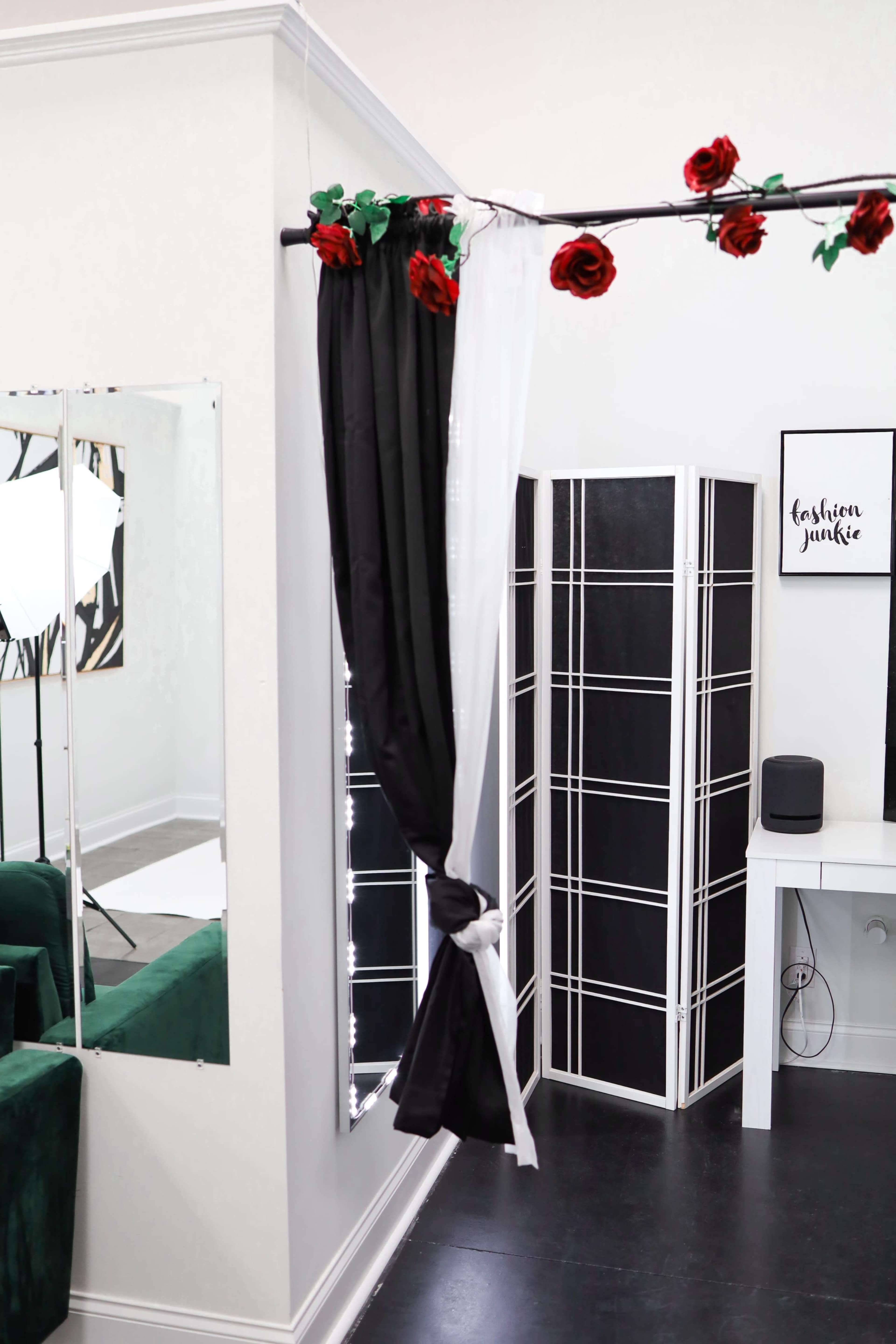 The image shows a room corner with a black curtain tied back, a folding screen, and a white desk, accented by red roses above.