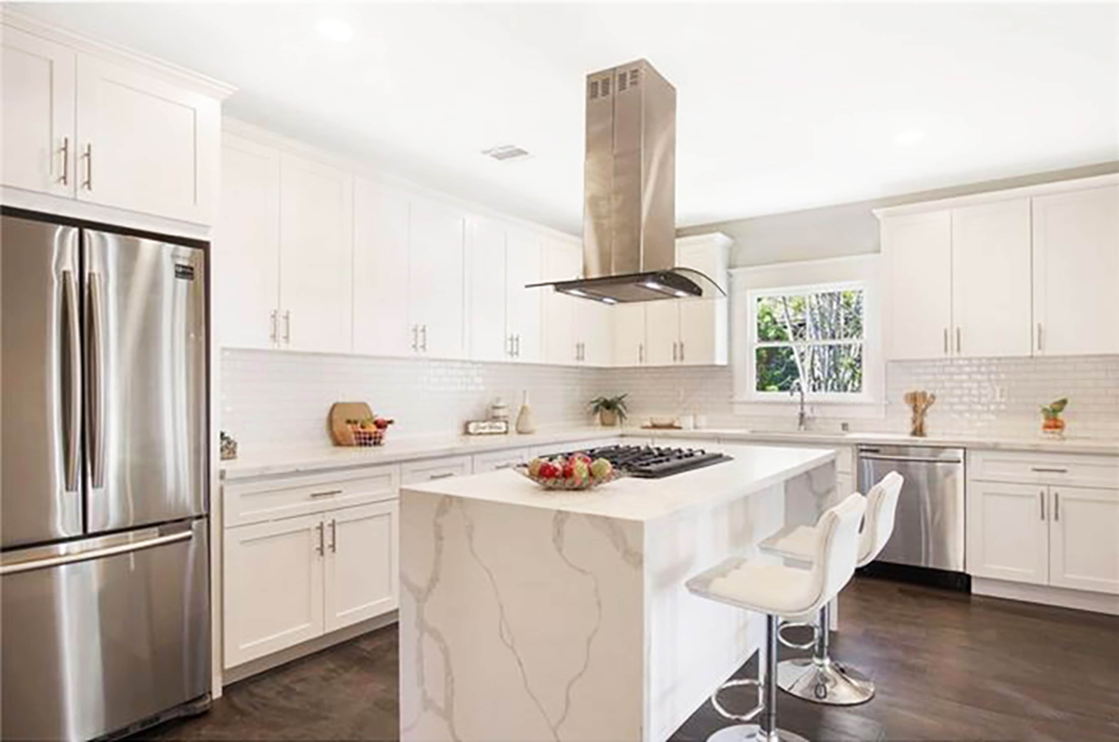 A modern kitchen with white cabinetry, a marble island, stainless steel appliances, and a sleek vent hood over the cooktop.