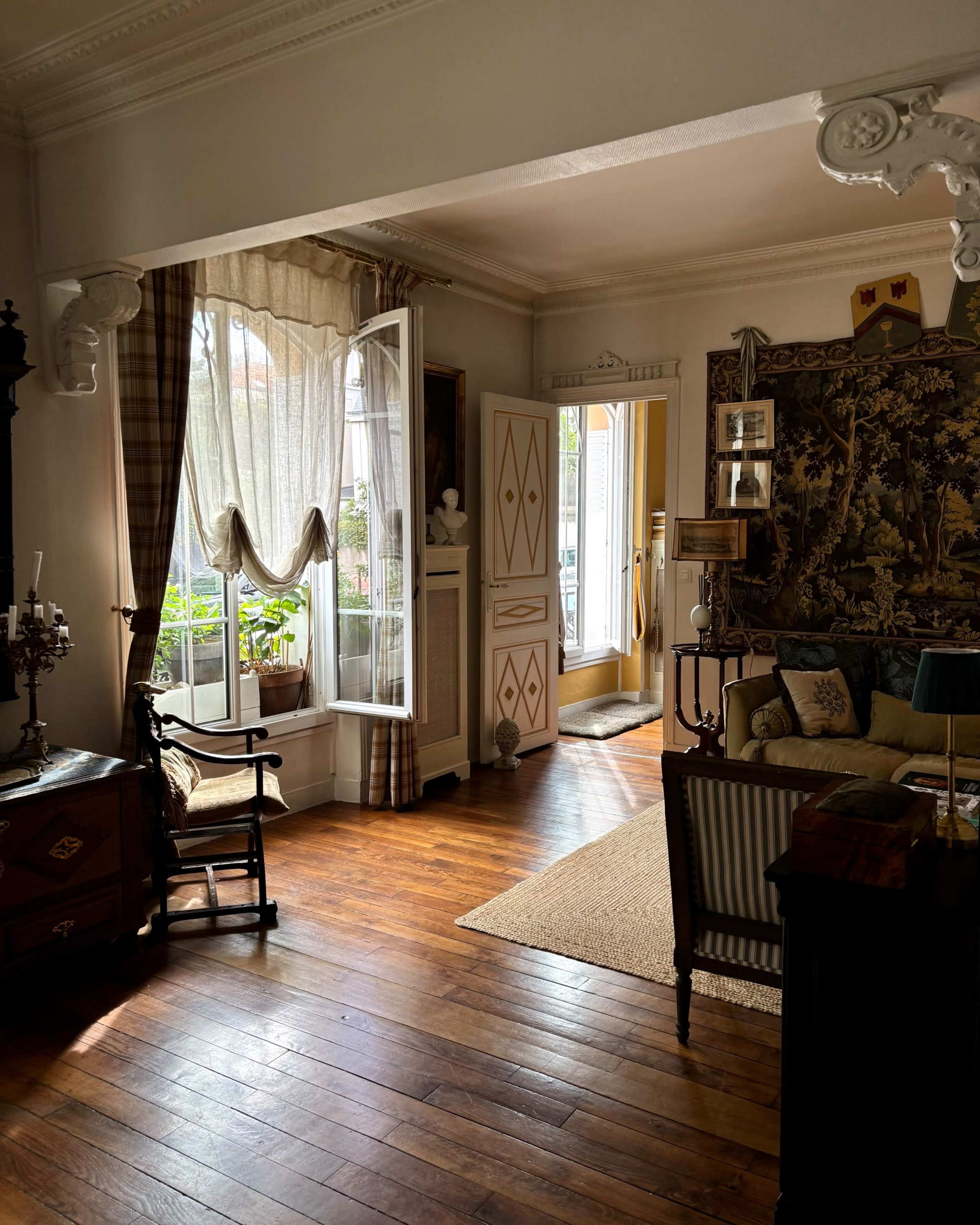 The image shows a well-lit living room with wooden floors, featuring a large window, a comfortable seating area, and decorative elements like a tapestry and various furniture pieces.