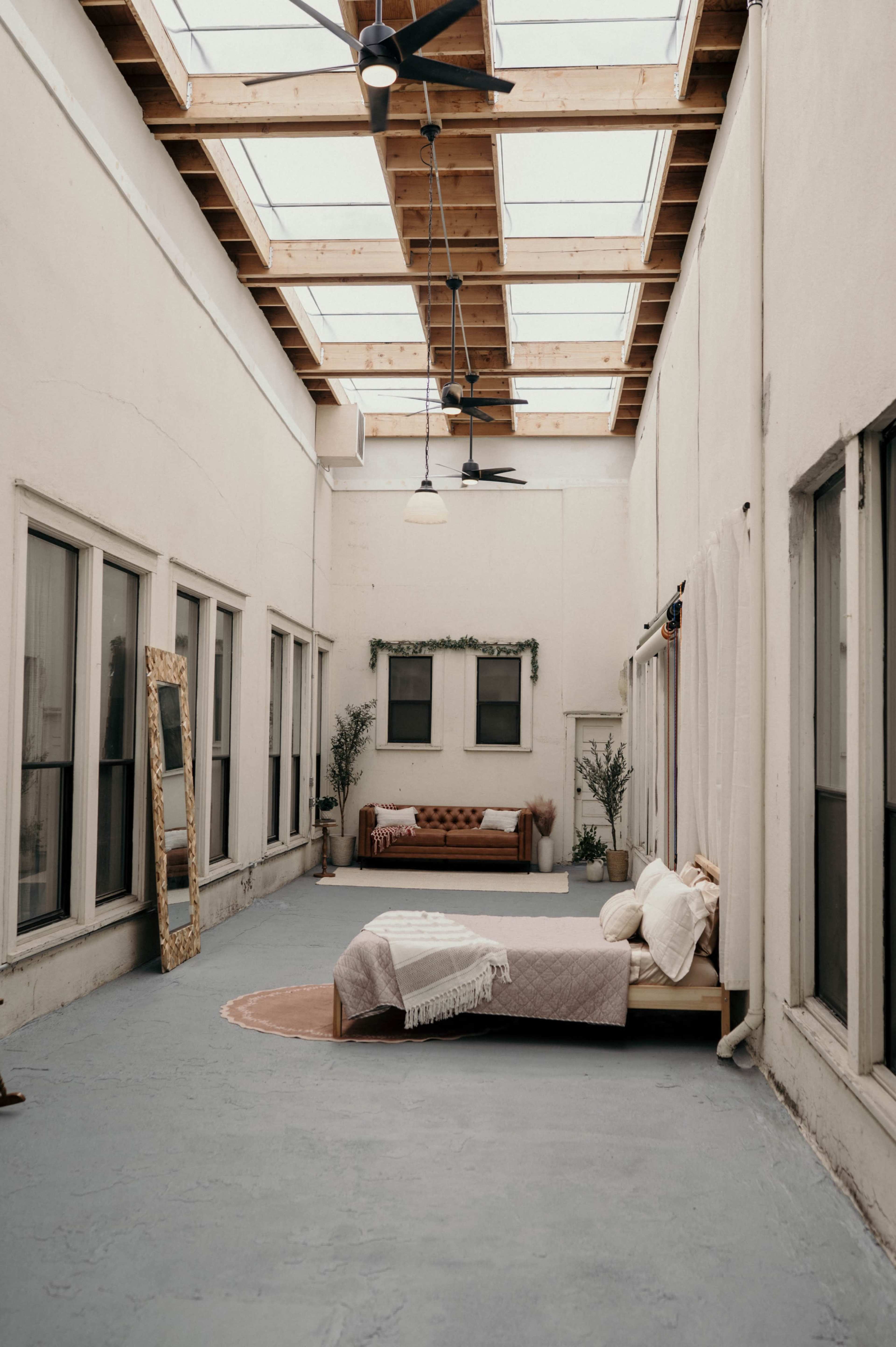 The image shows a spacious, bright interior room featuring a bed, a couch, a large mirror, and windows along the walls.