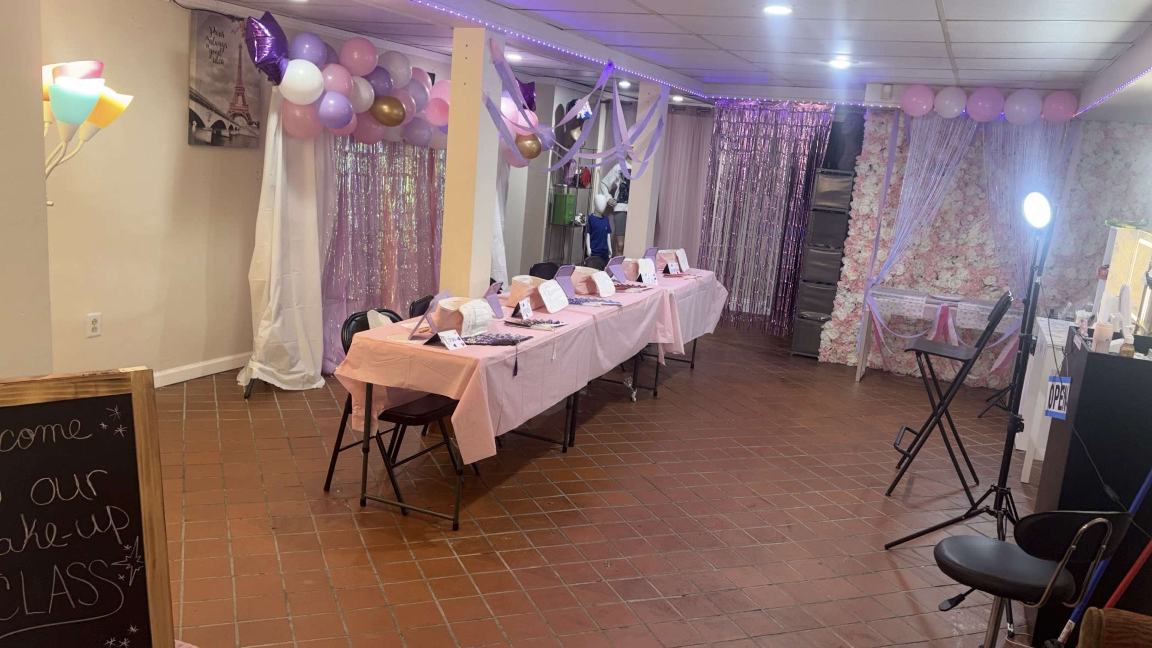The image shows a decorated indoor space set up for a makeup class, featuring several tables covered with pink linens, chairs, and colorful balloons arranged along the walls.
