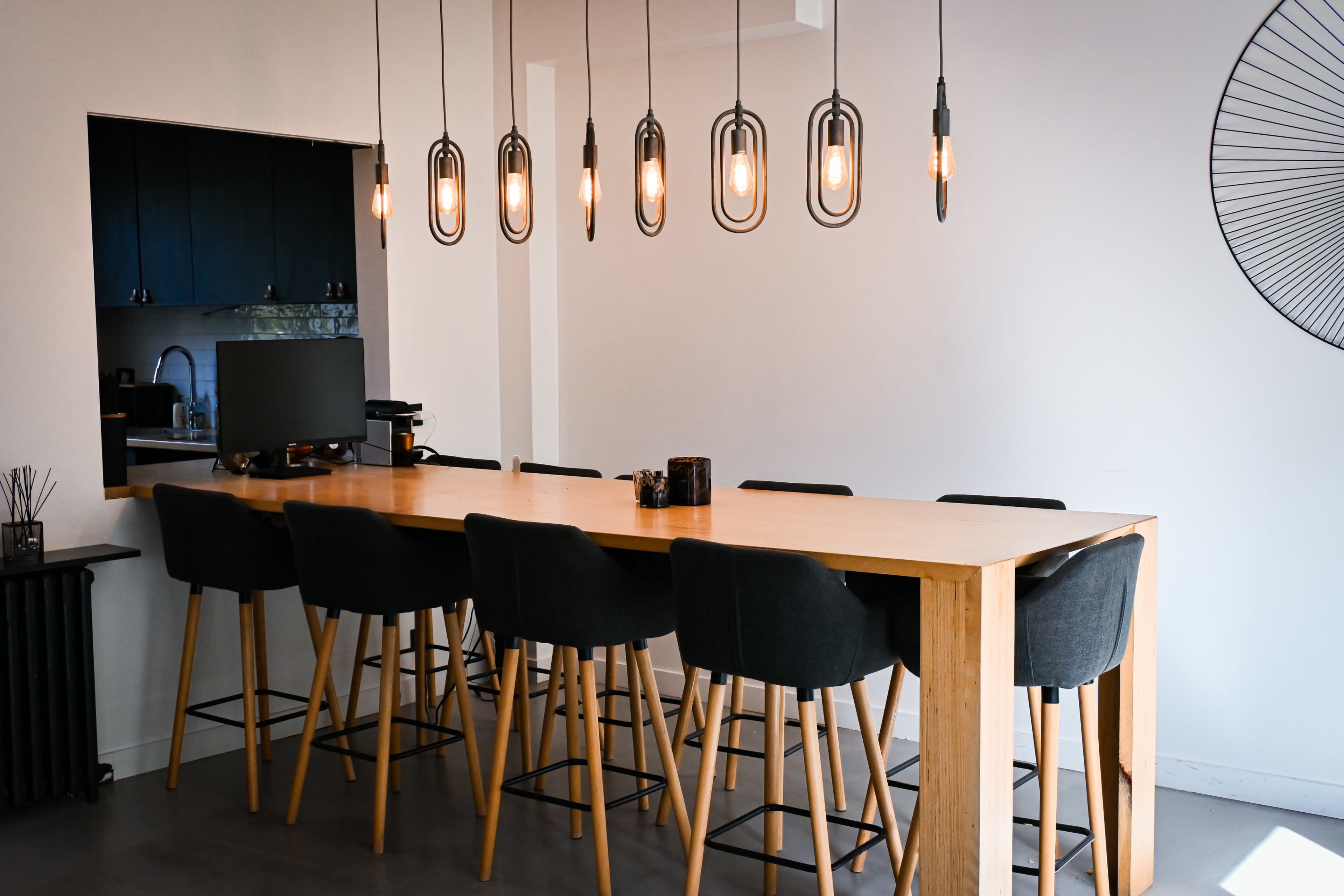 The image shows a modern kitchen with a long wooden dining table surrounded by high-backed chairs and stylish pendant lights overhead.