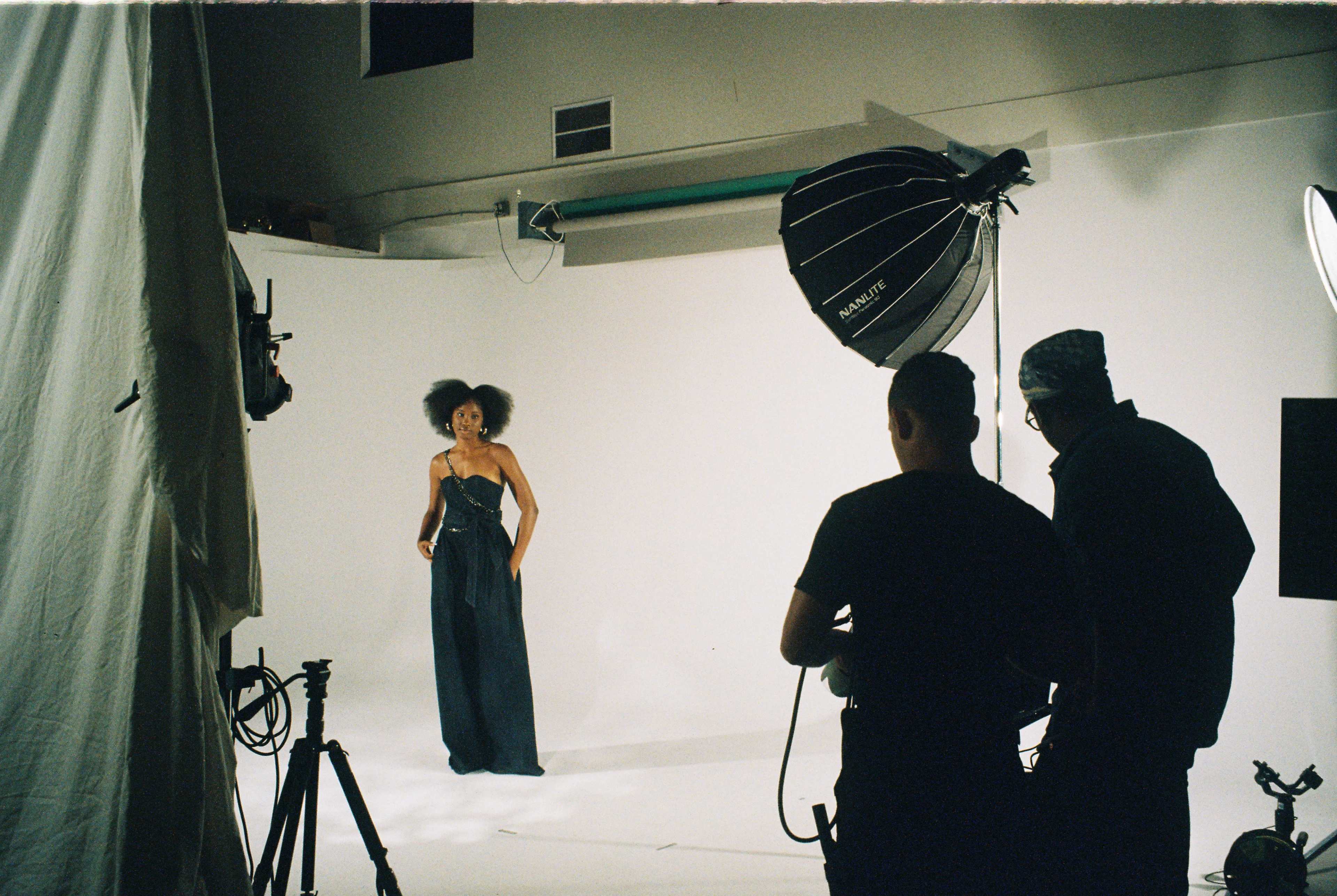 A model stands in front of a plain white backdrop while two crew members adjust lighting during a photoshoot.