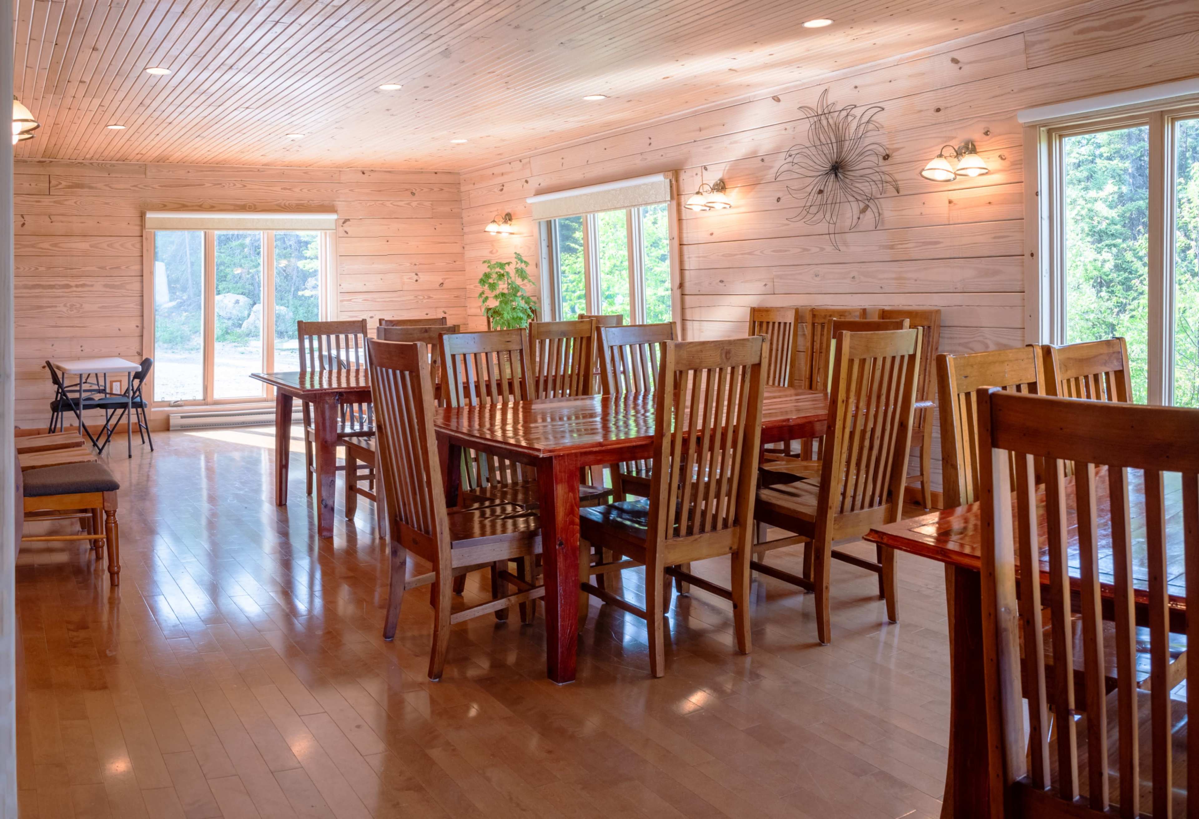 A spacious dining area features wooden tables and chairs arranged neatly along a light-colored wooden floor, with large windows allowing natural light to enter.