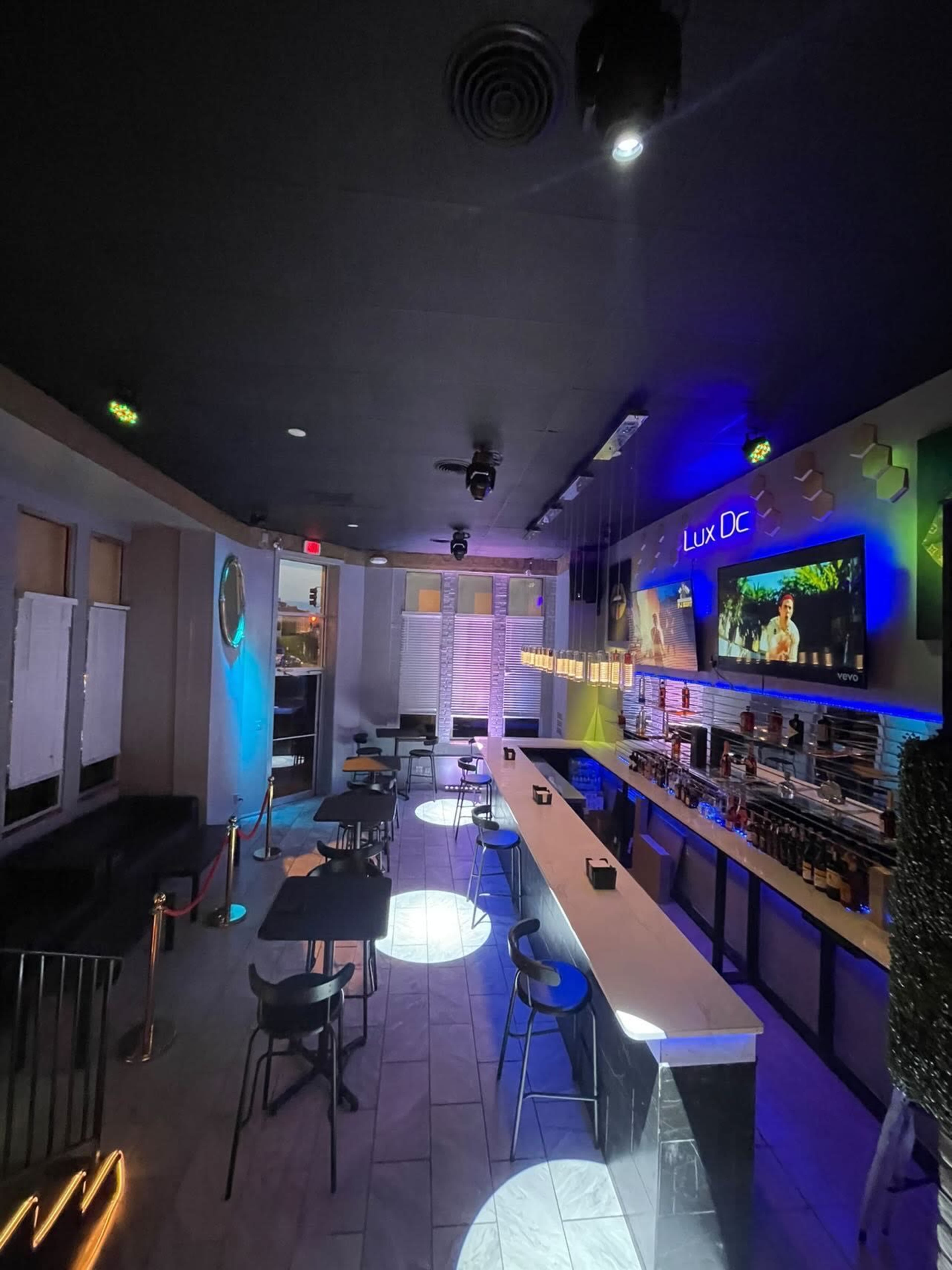 The image shows a modern bar with a long counter, chairs, and colorful lighting in a dimly lit space.