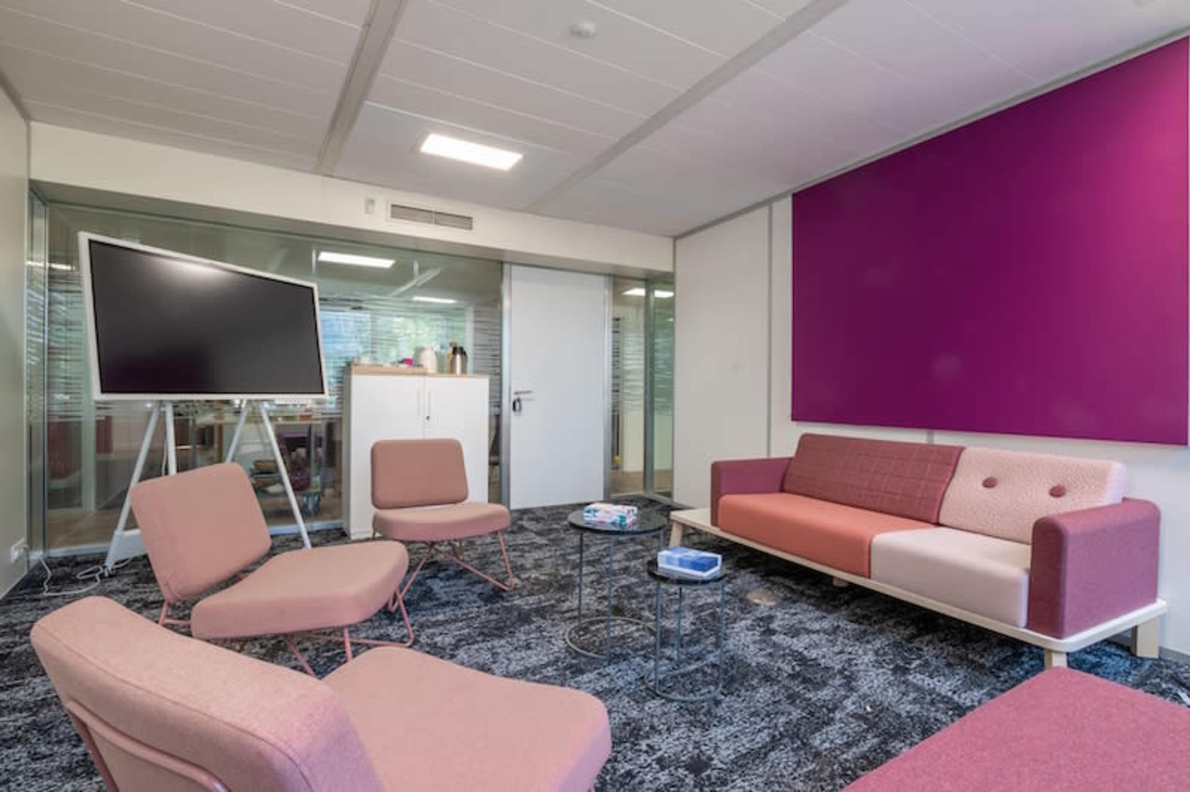 A modern meeting room with pink and gray seating, a large TV on a stand, and a purple accent wall.
