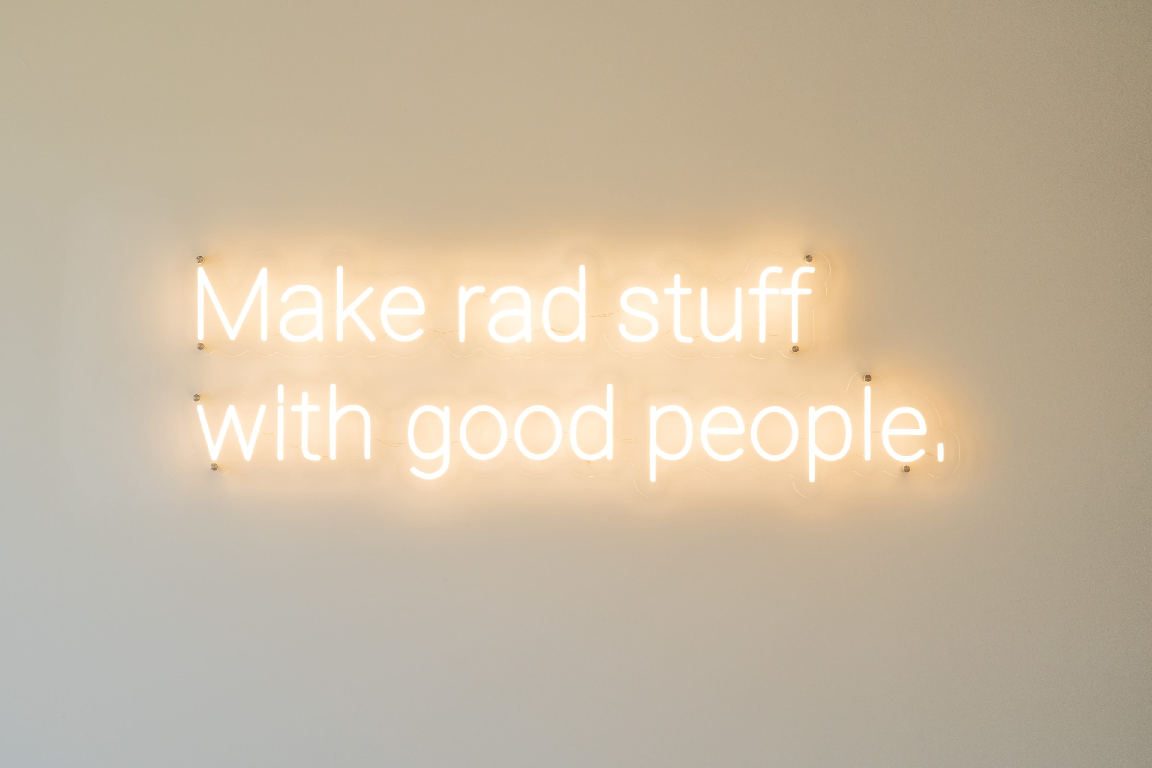 A neon sign on a wall reads, "Make rad stuff with good people."