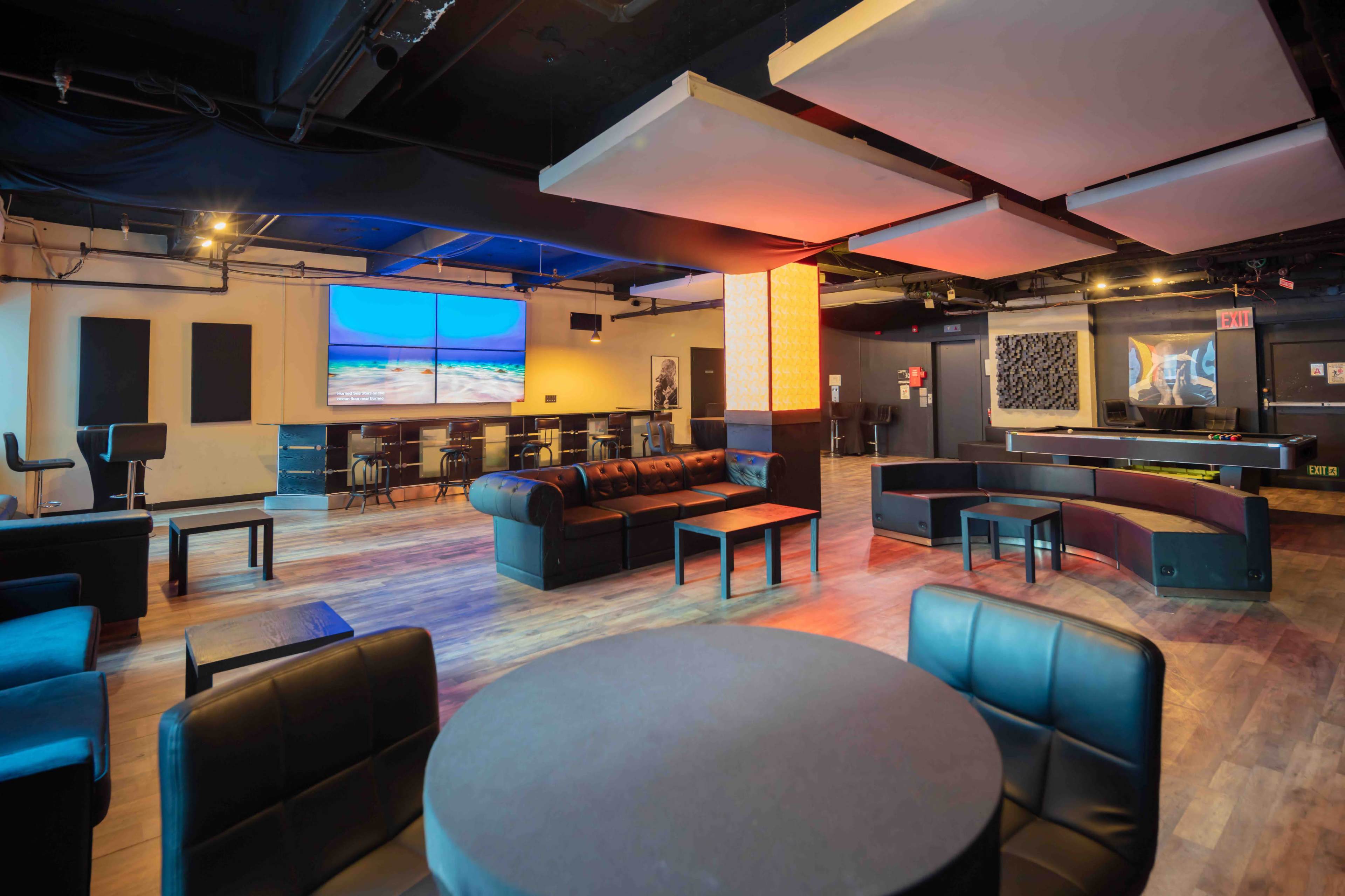 The image shows a spacious lounge area with black leather seating, flat screen televisions, and a wooden floor, designed for social gatherings.
