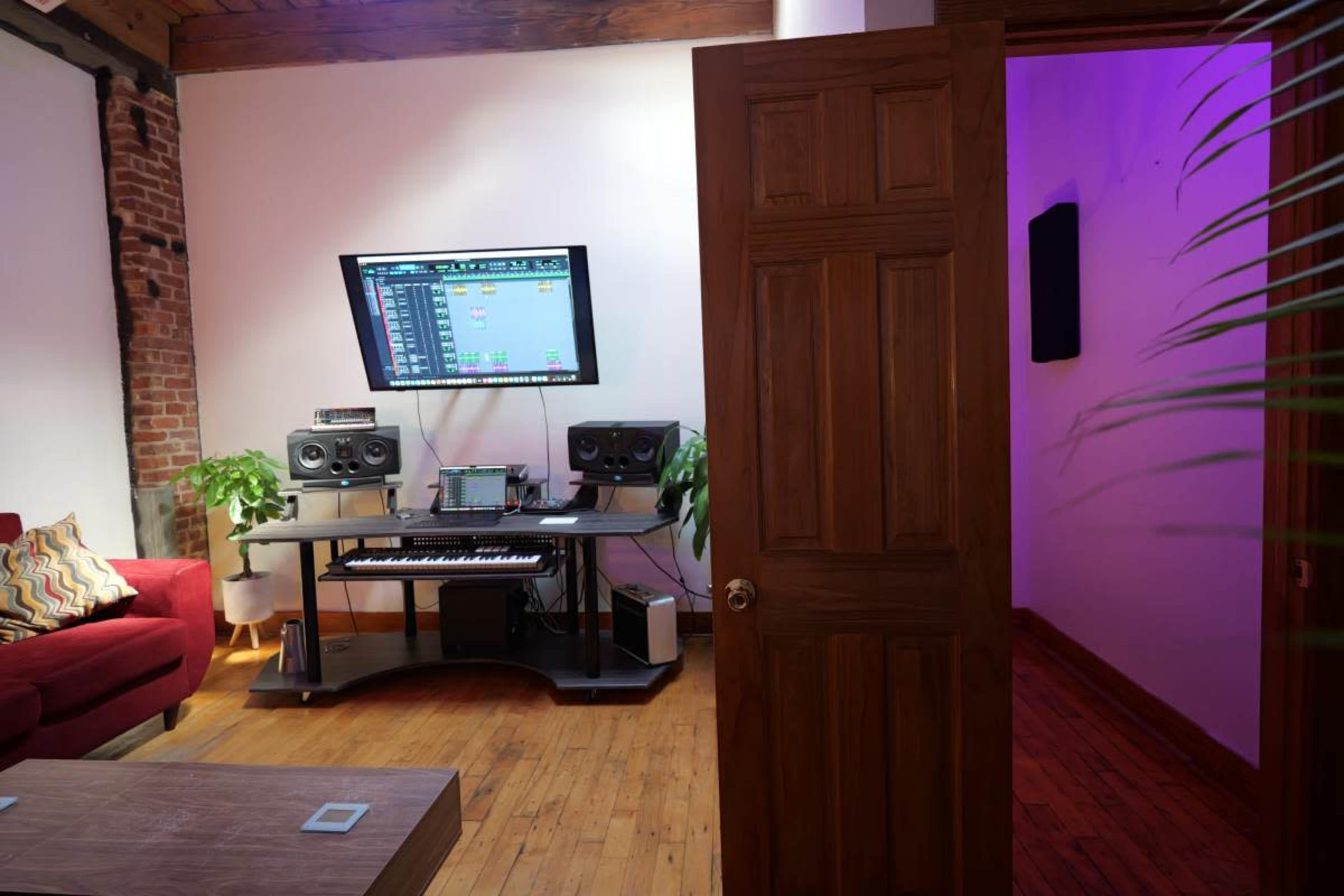 The image shows a home music studio featuring a large screen displaying audio software, a keyboard, speakers, and plants, with a door leading to a purple-lit room in the background.