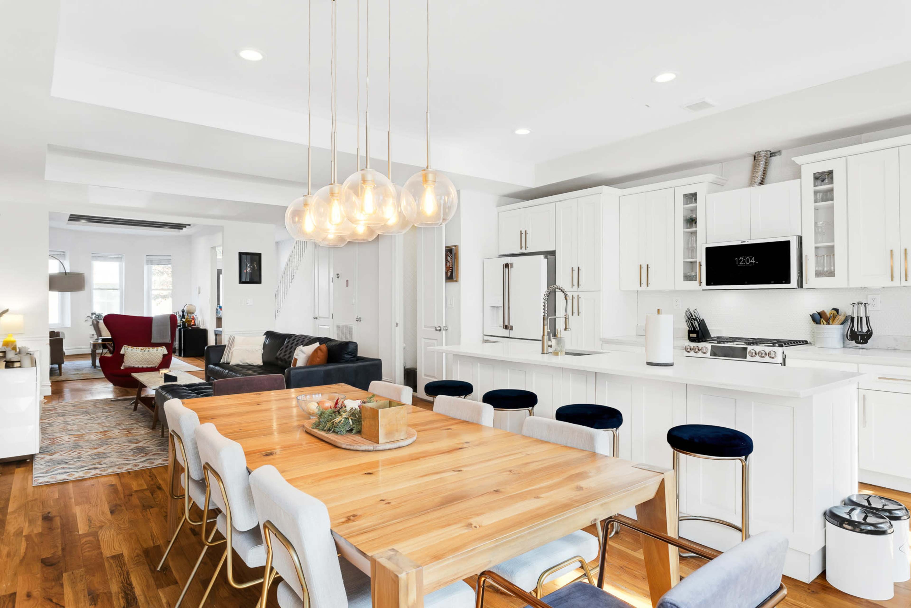 A modern kitchen and dining area features a long wooden table surrounded by upholstered chairs, with pendant lights hanging above and a spacious living area visible in the background.
