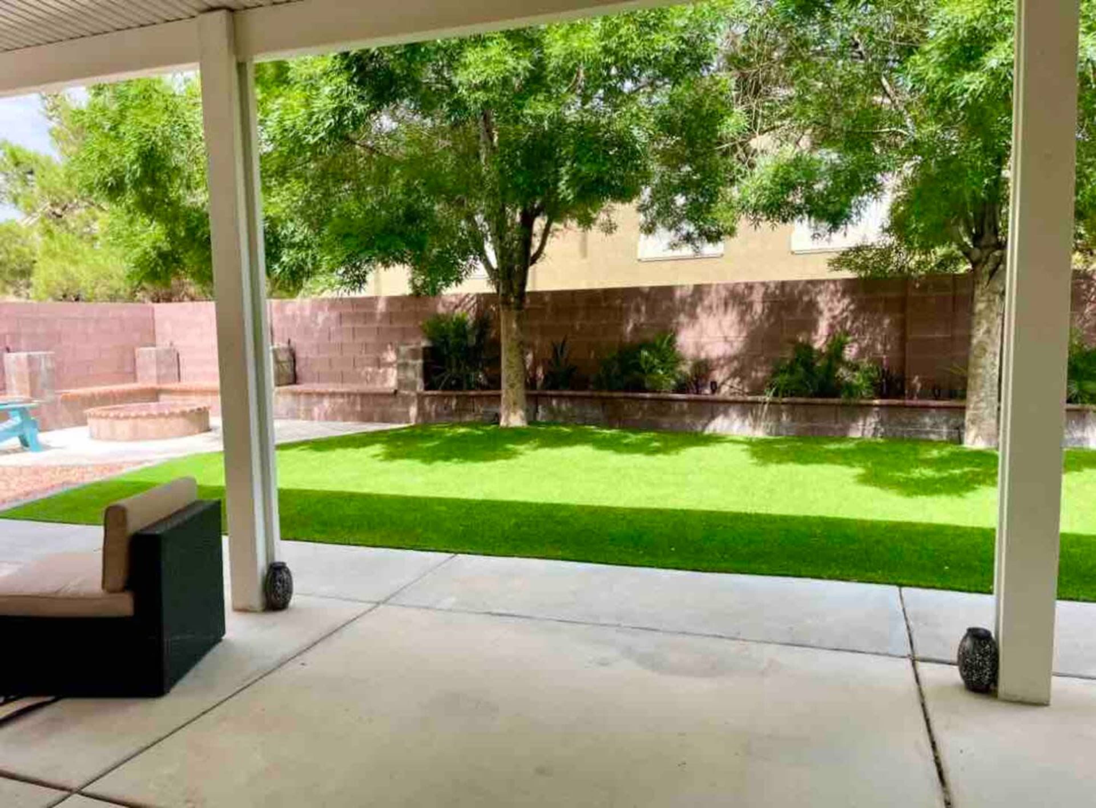 The image shows a covered patio viewing a green lawn surrounded by trees and a stone wall in the background.