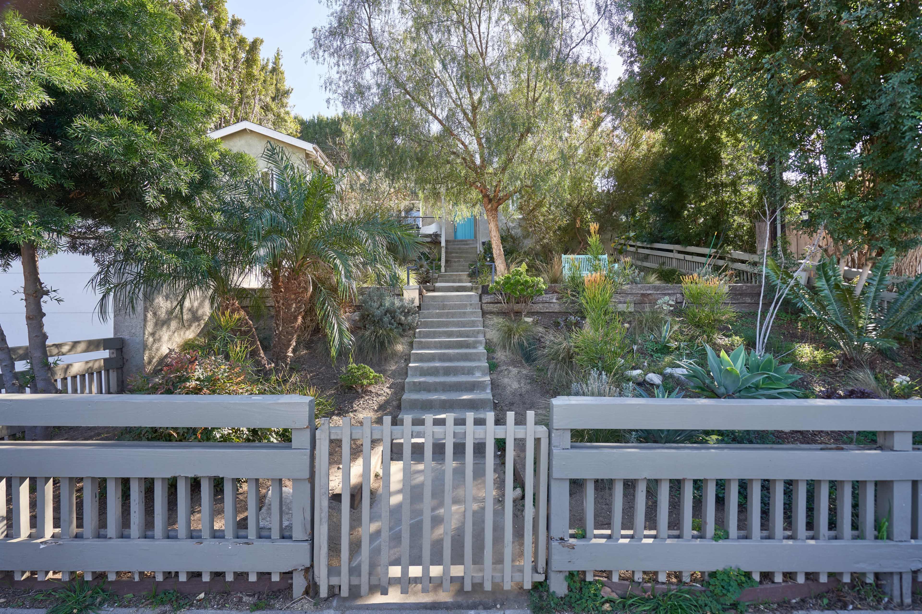A gray picket fence encloses a landscaped front yard with various plants and a set of stone steps leading to a house.