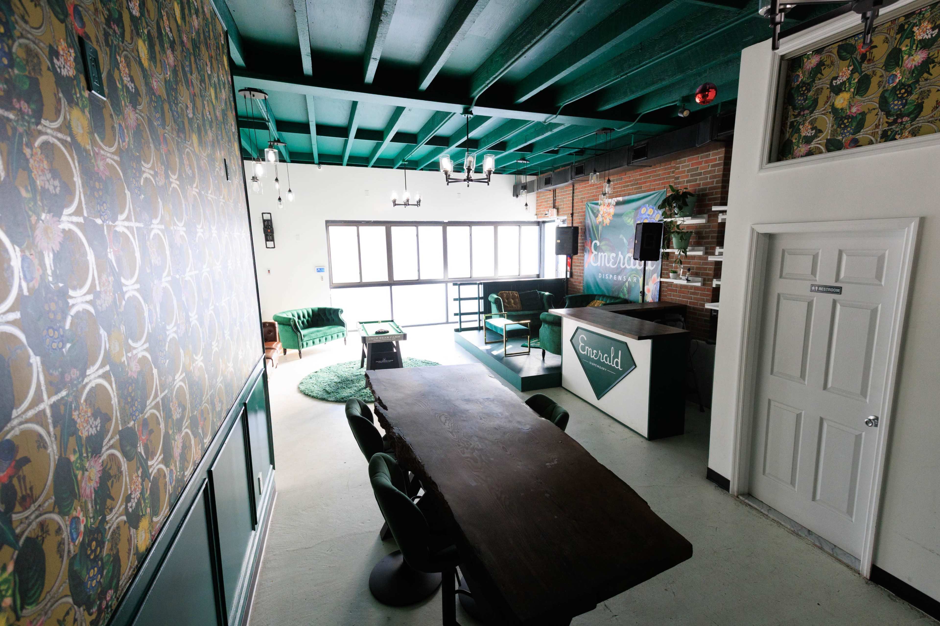The image shows a modern interior space with green-painted beams, a long wooden table, and a bar area featuring a sign that reads "Emerald."