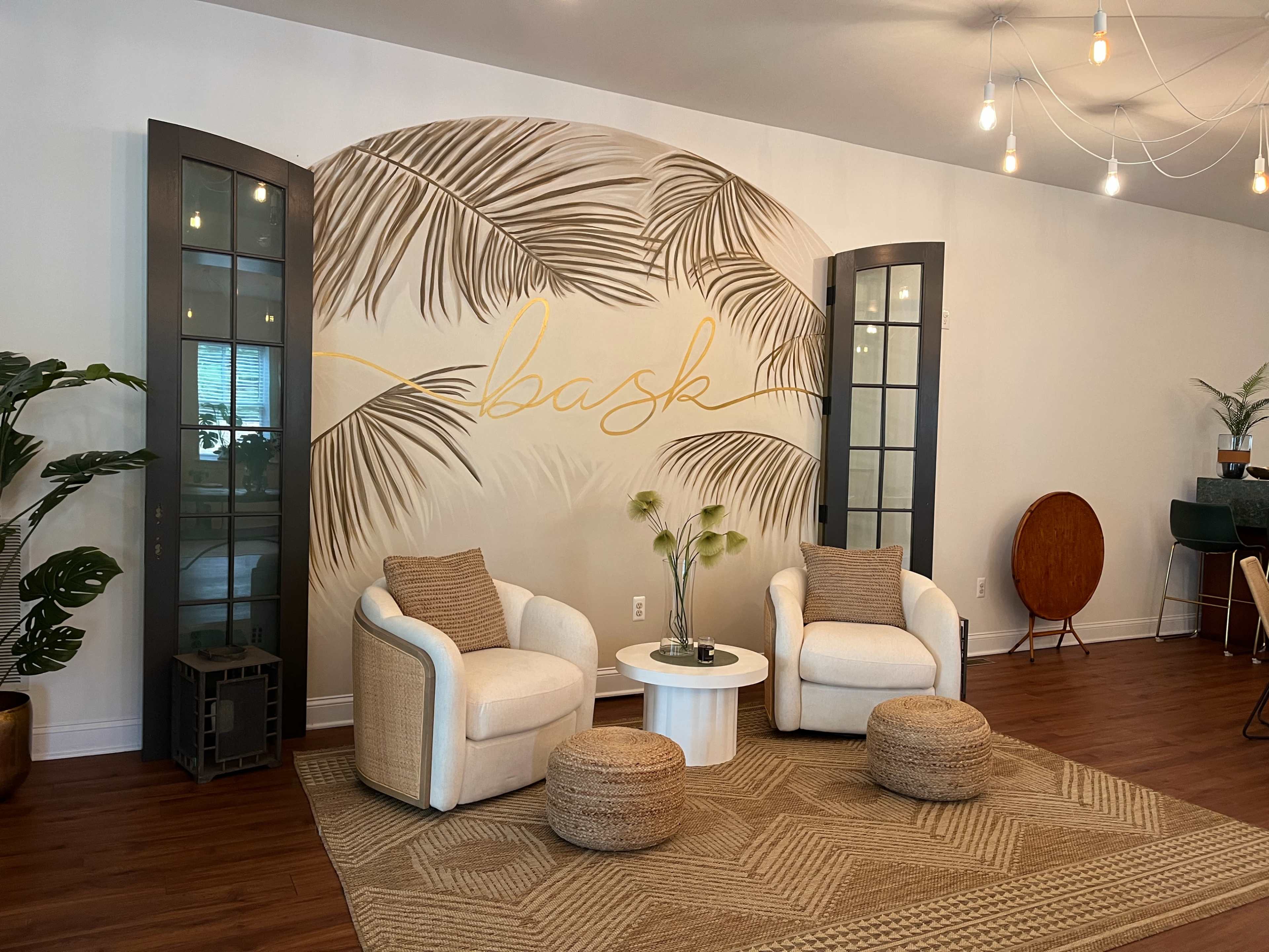The image shows a cozy sitting area with two white armchairs, a small round table, and decorative poufs on a rug, complemented by a large wall mural featuring palm leaves and the word "bask."