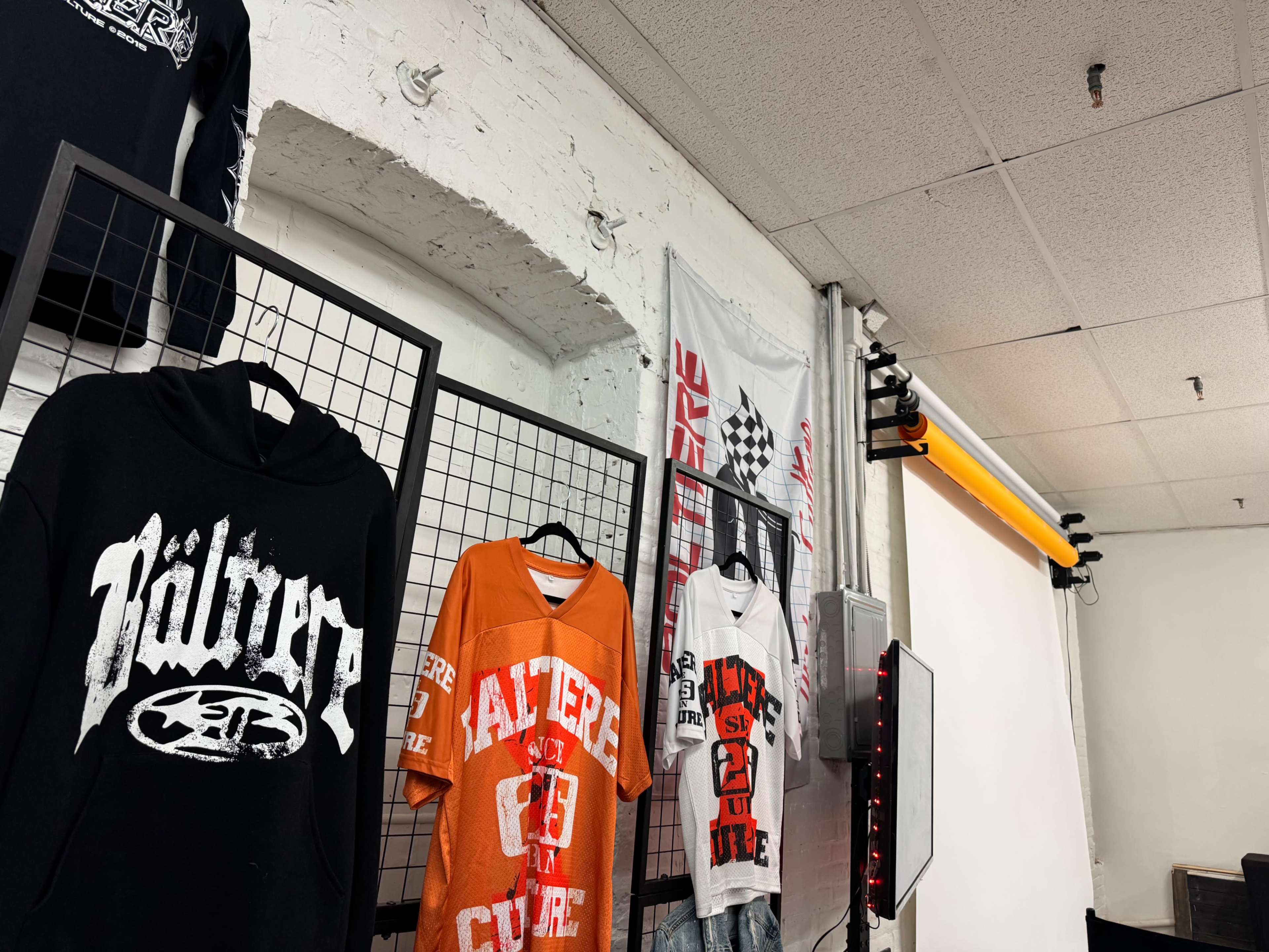 The image features a wall display of various graphic t-shirts arranged on black grid panels, with a backdrop of white fabric and a yellow lighting strip.
