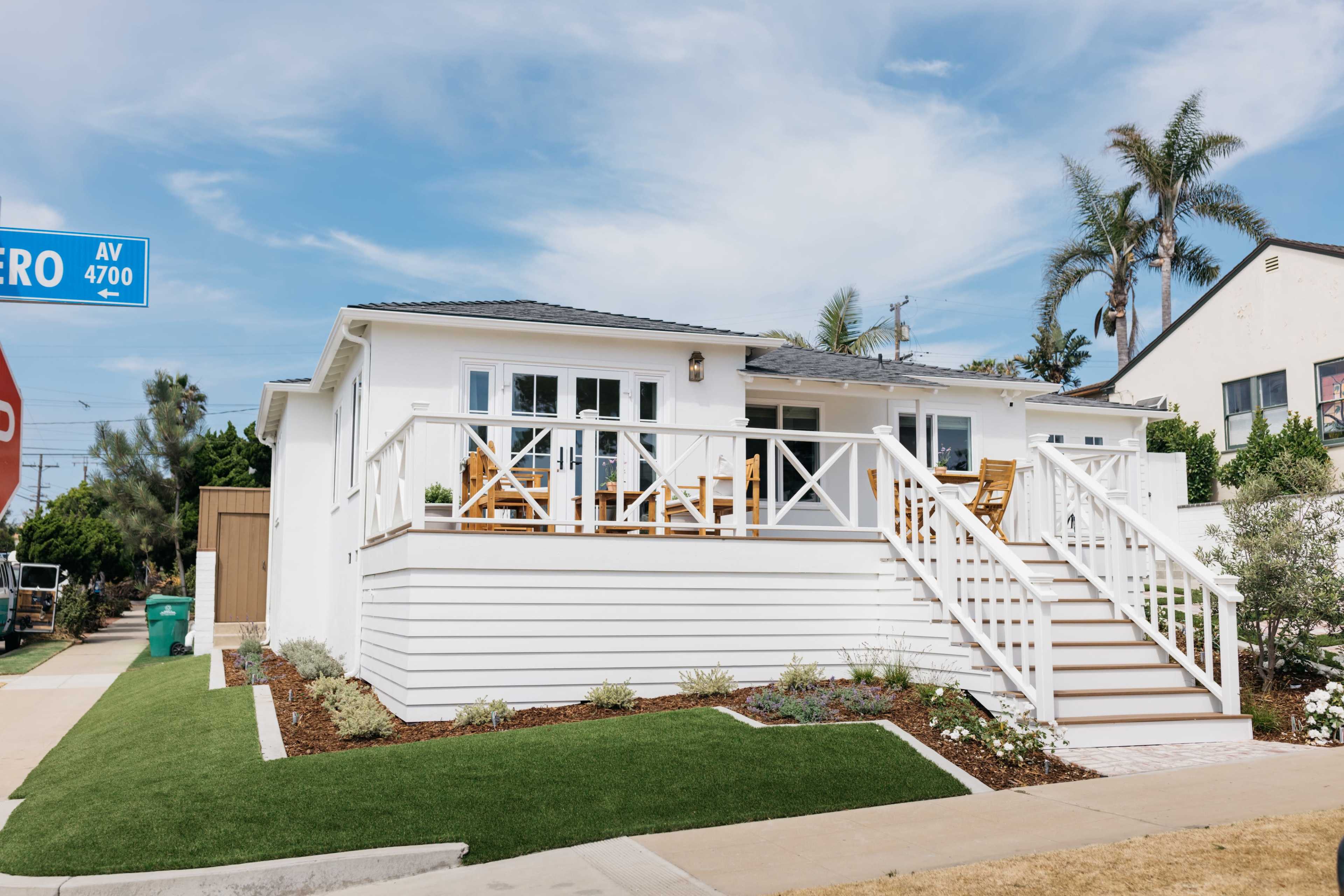 A single-story white house with a porch and staircase is situated on a corner lot, surrounded by landscaped greenery and palm trees.