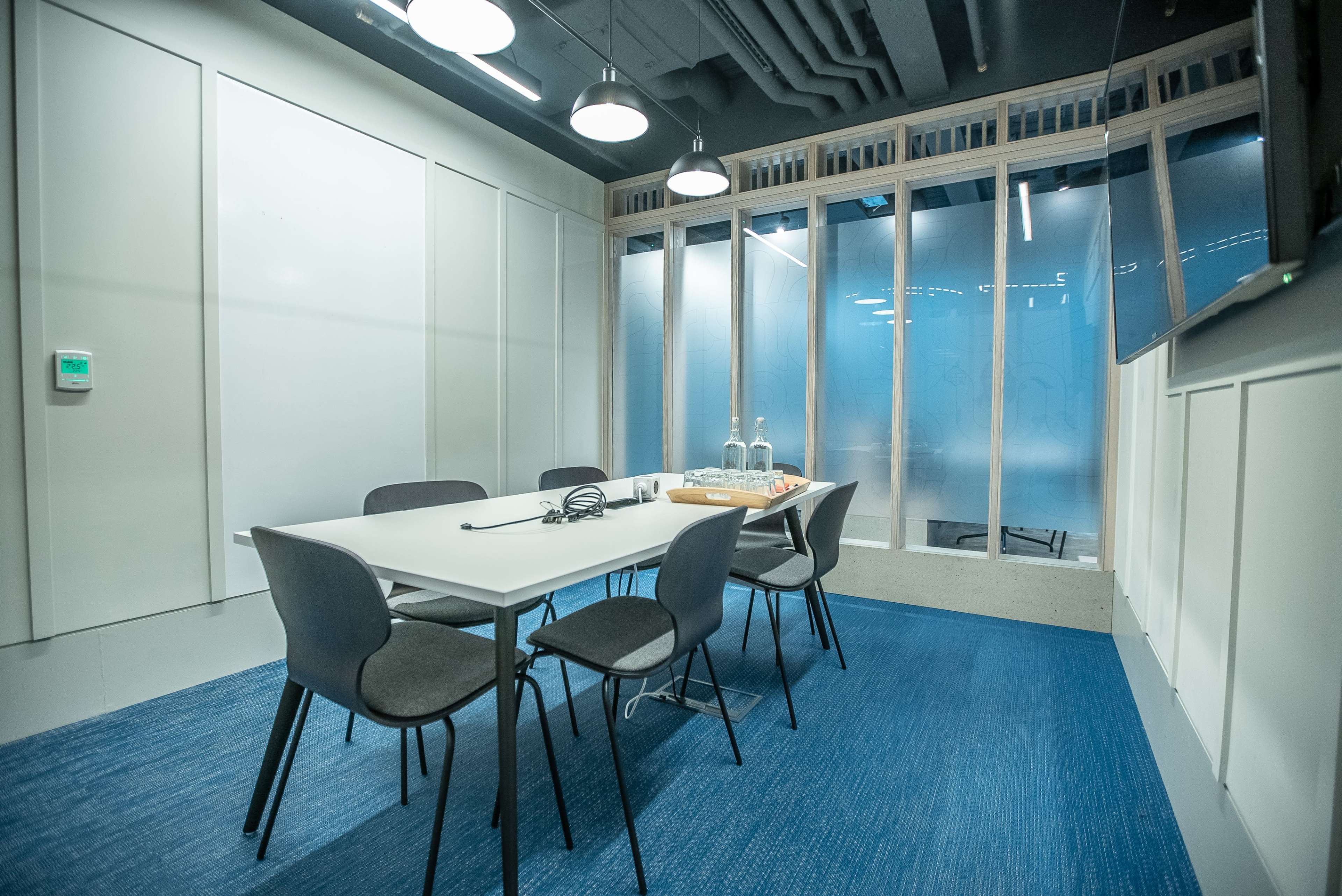 A conference room features a rectangular table surrounded by chairs, with glass walls and a blue carpet.