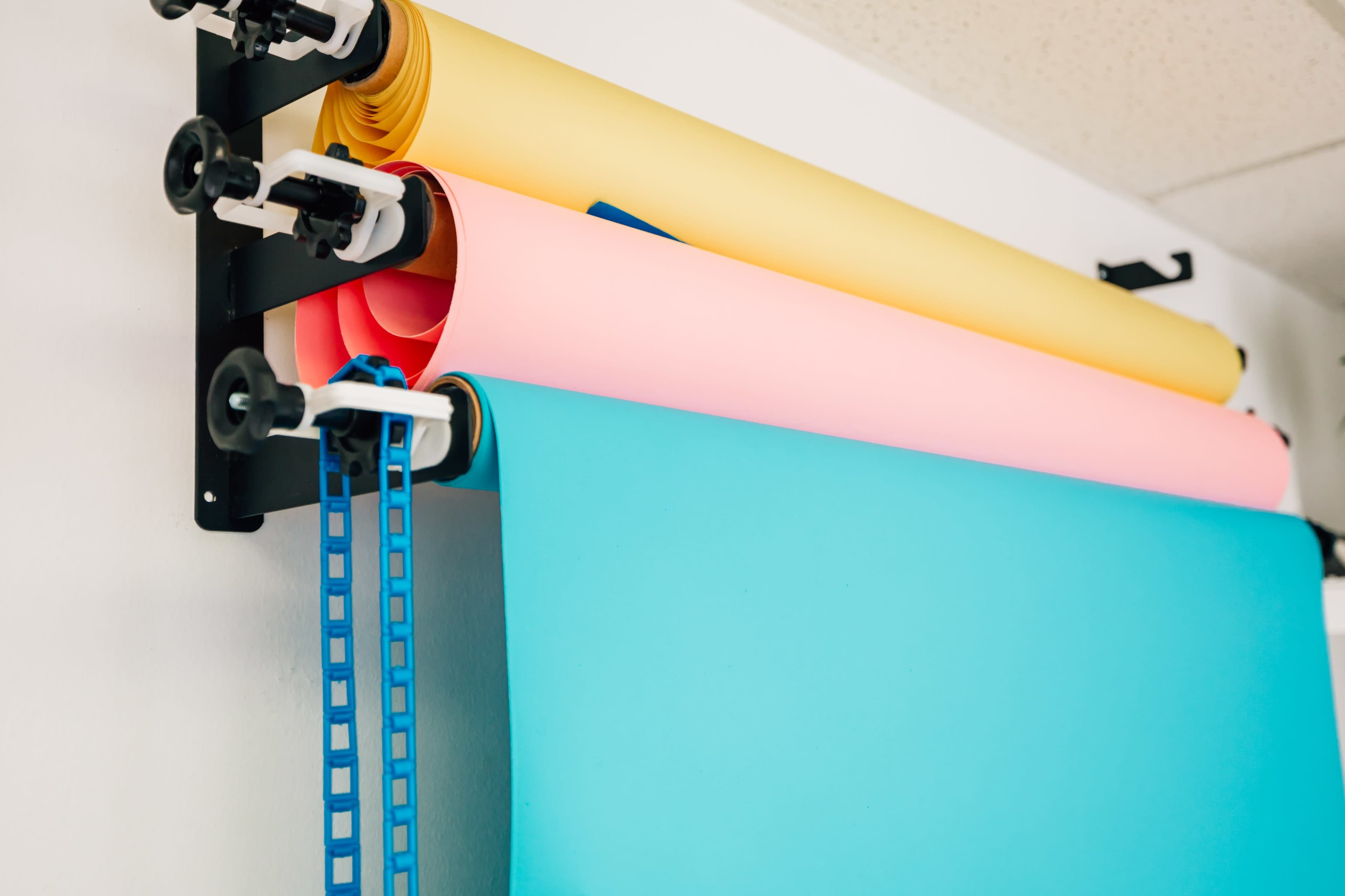 The image shows three rolls of colored paper—yellow, pink, and blue—mounted on a wall rack.