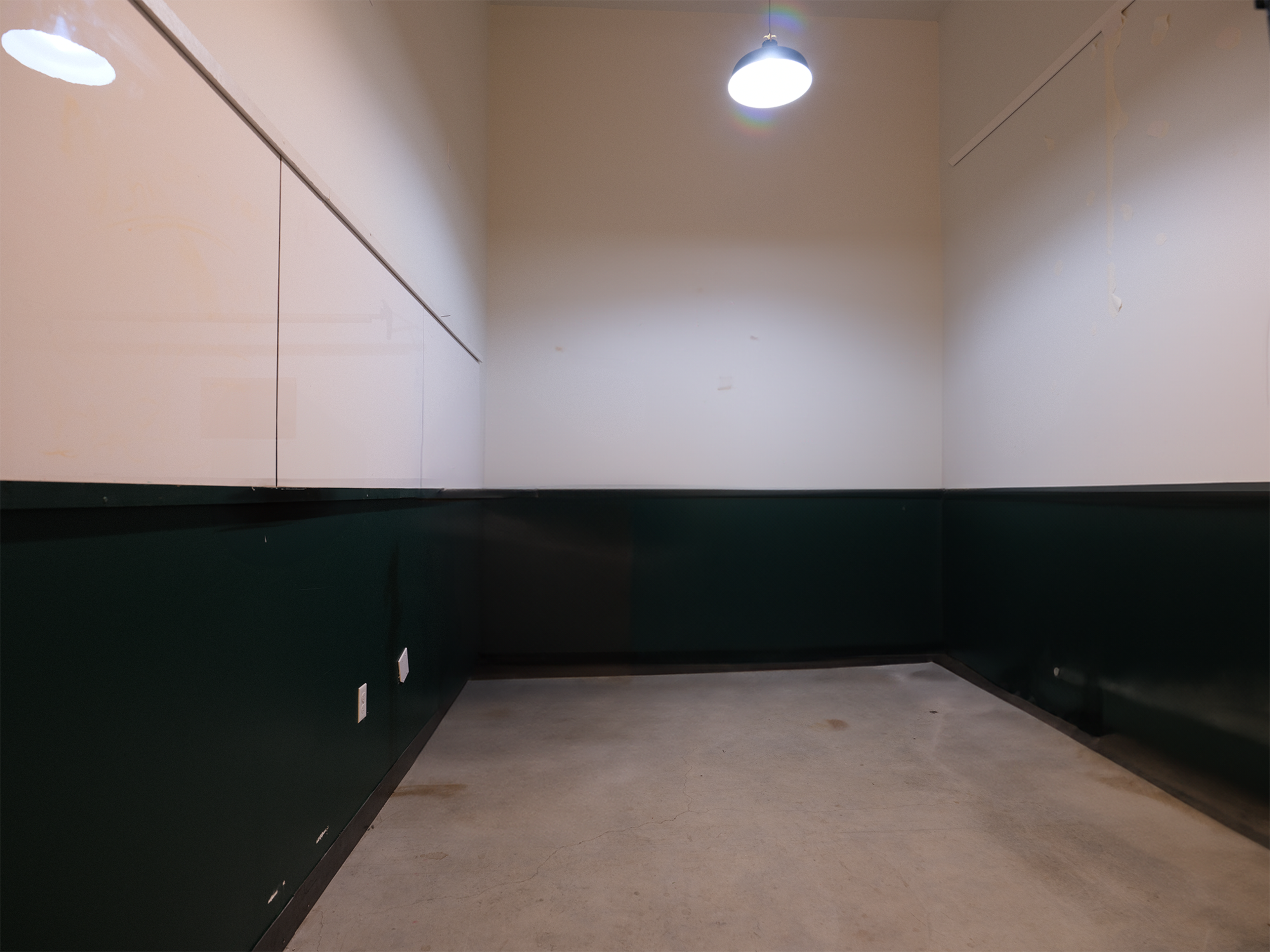 The image shows a sparsely furnished room featuring plain white walls at the top and dark green walls below, with a single overhead light illuminating the space.