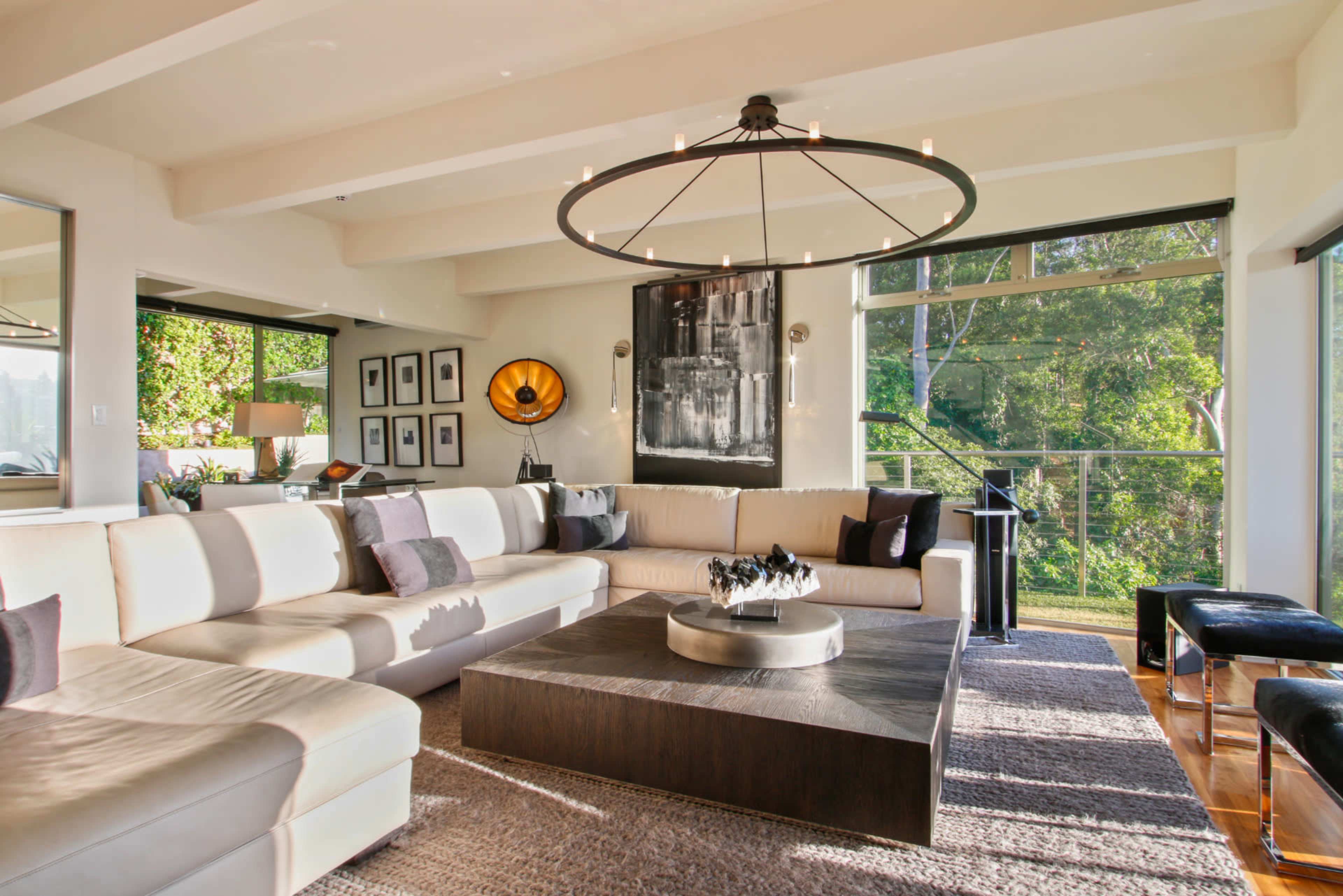The image shows a modern living room featuring a large sectional sofa, a circular chandelier, and a coffee table with a fire pit, surrounded by large windows that offer a view of greenery outside.