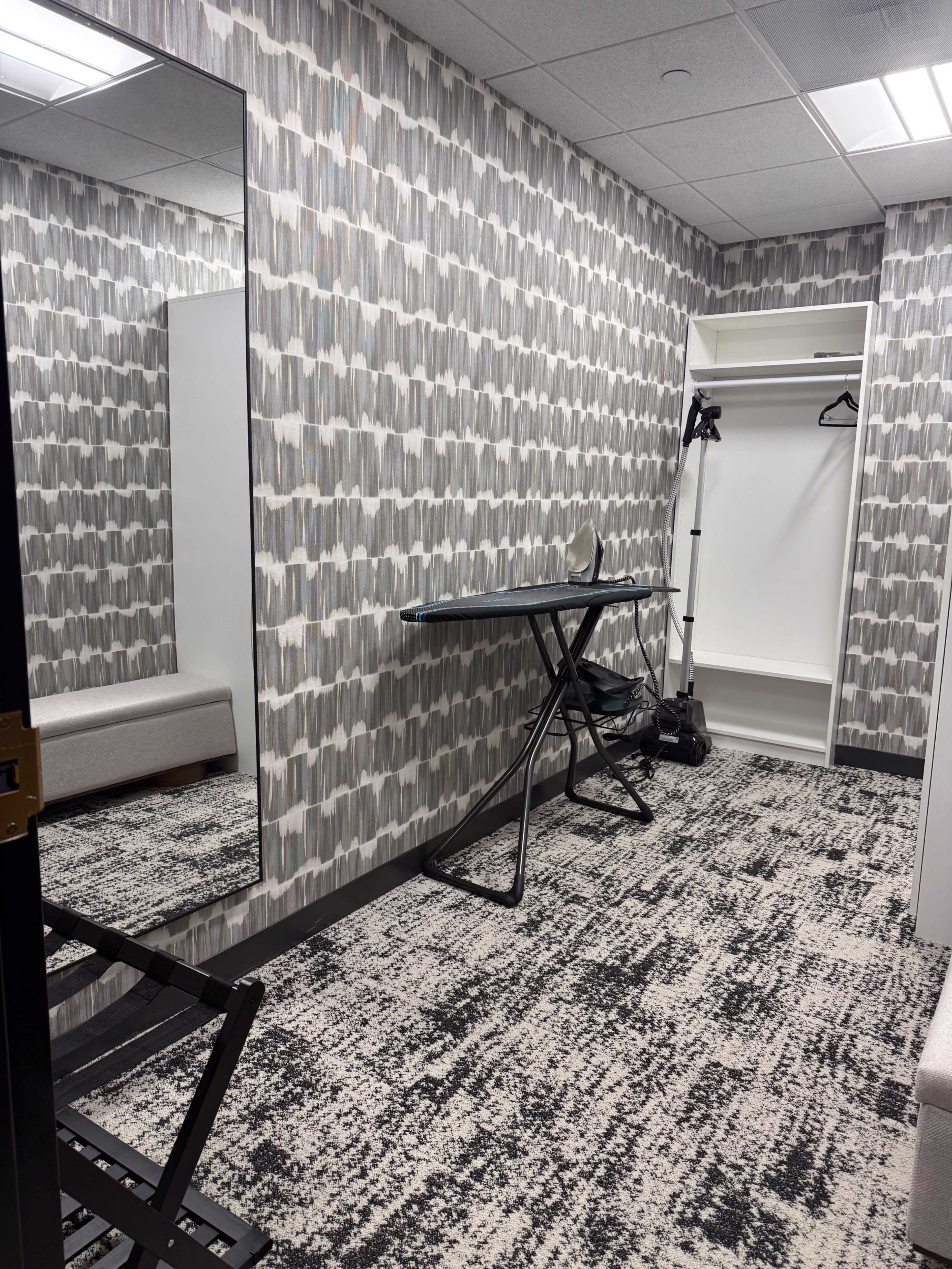 The image shows a small, well-lit dressing room featuring a mirror, a garment rack, an ironing board, and a patterned wallpaper.