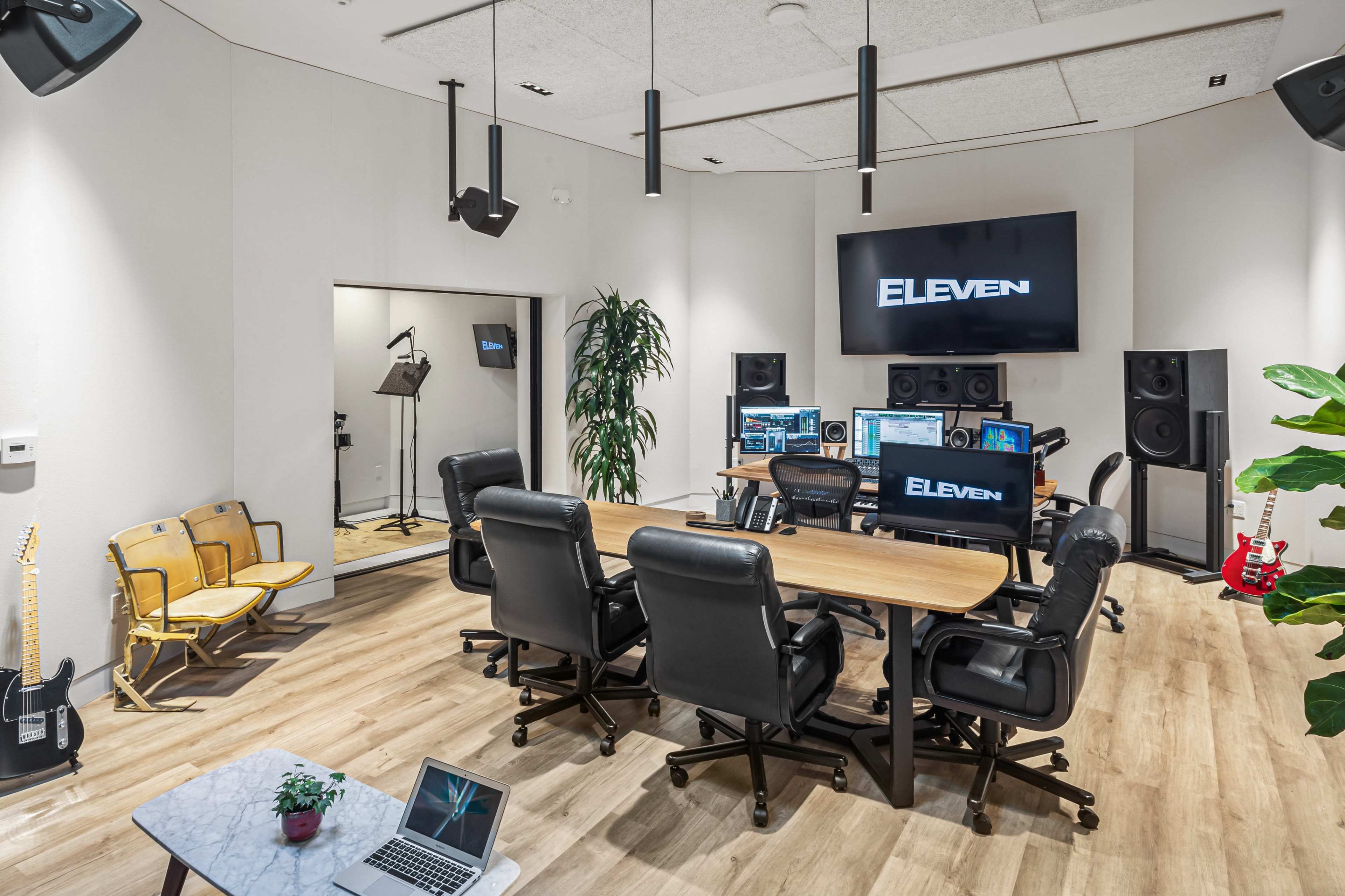 A modern recording studio equipped with a large screen displaying "ELEven," multiple computer workstations, audio monitors, and a small seating area.