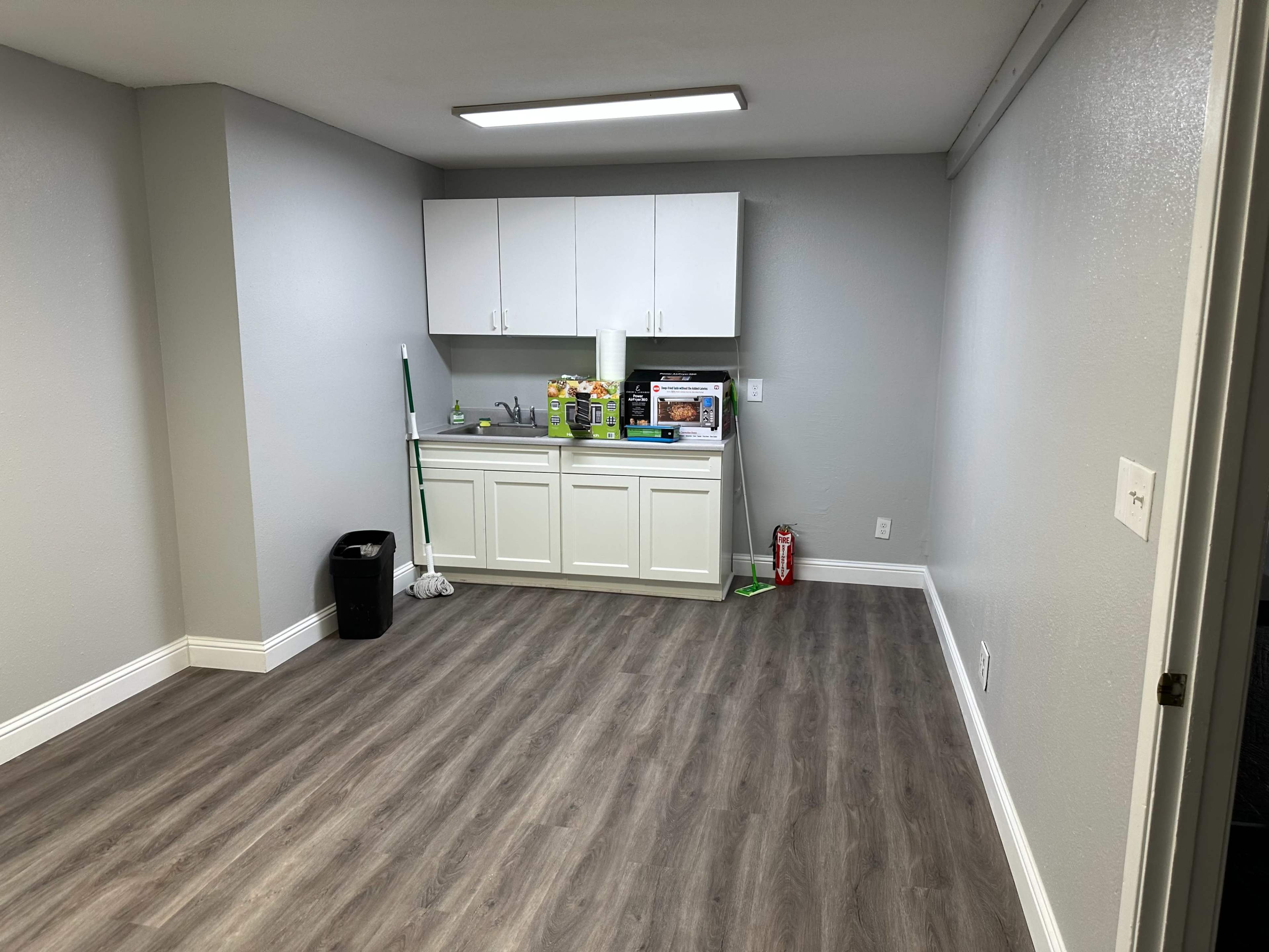 A clean, empty room with a kitchenette featuring white cabinets and a countertop, along with a mop, a trash can, and a fire extinguisher.