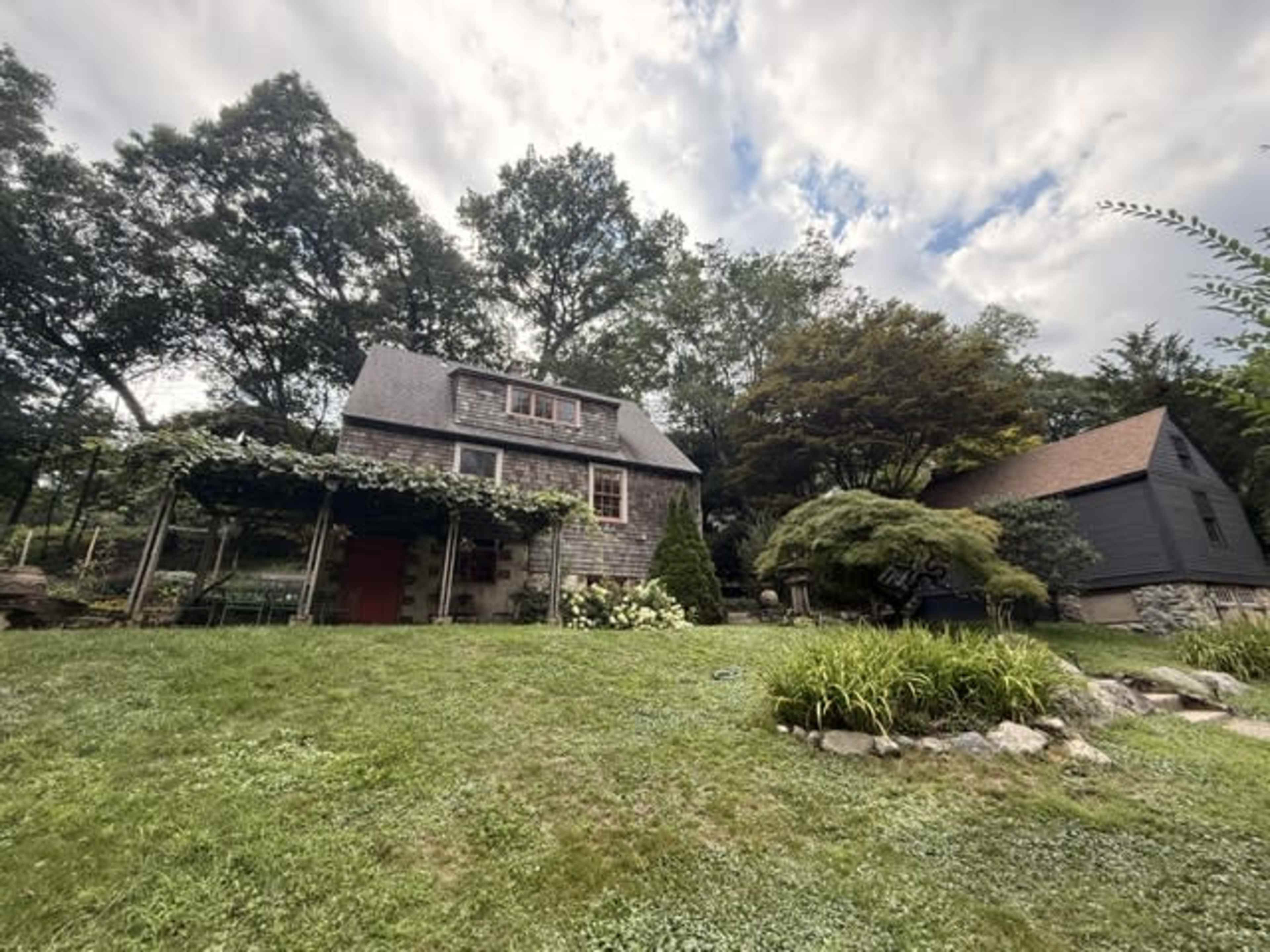 The image shows a two-story house with a stone exterior, surrounded by trees and a landscaped yard featuring green grass and various plants.