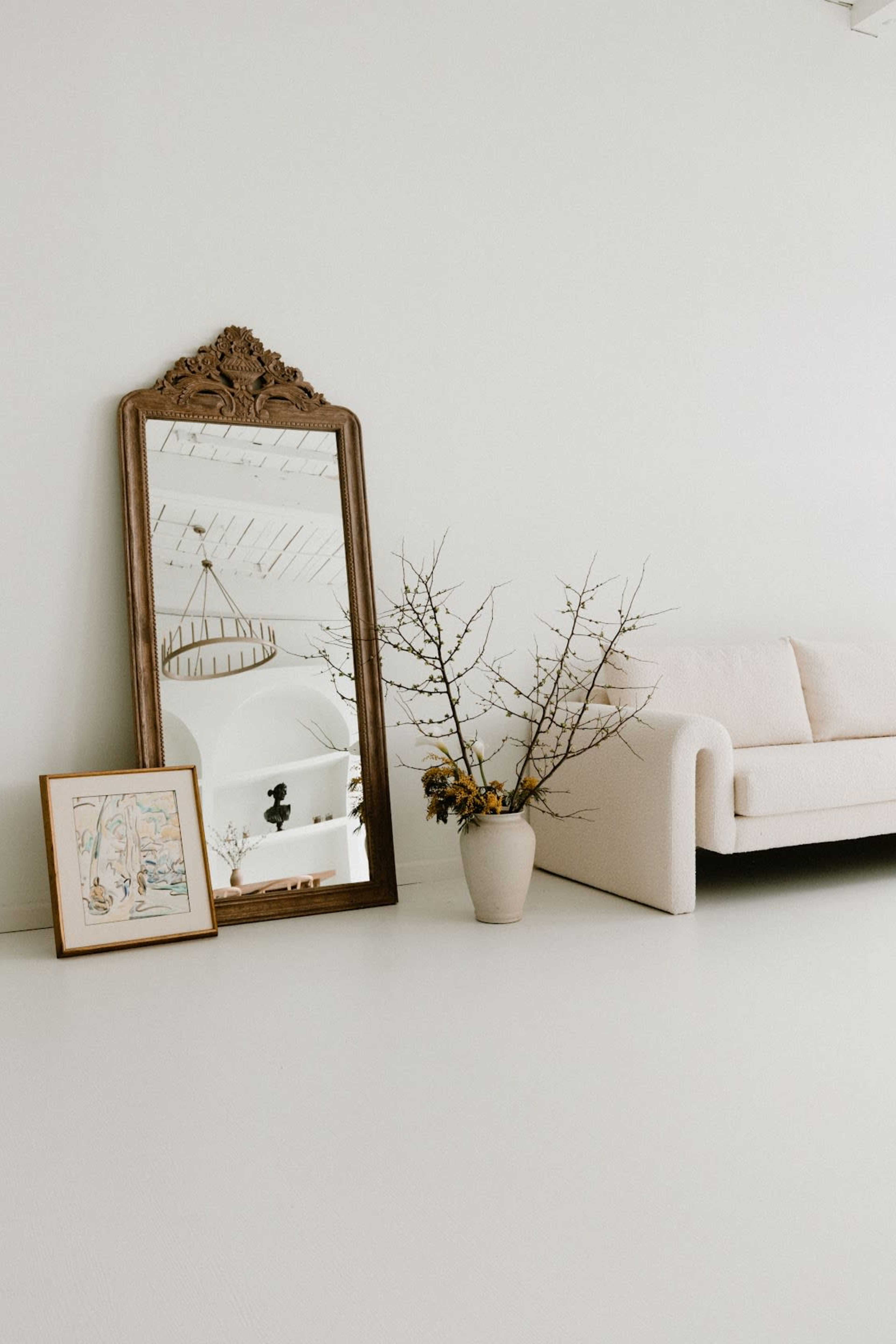 A large ornate mirror leans against a white wall beside a light-colored sofa, with a vase of branches and a framed artwork also on the floor.