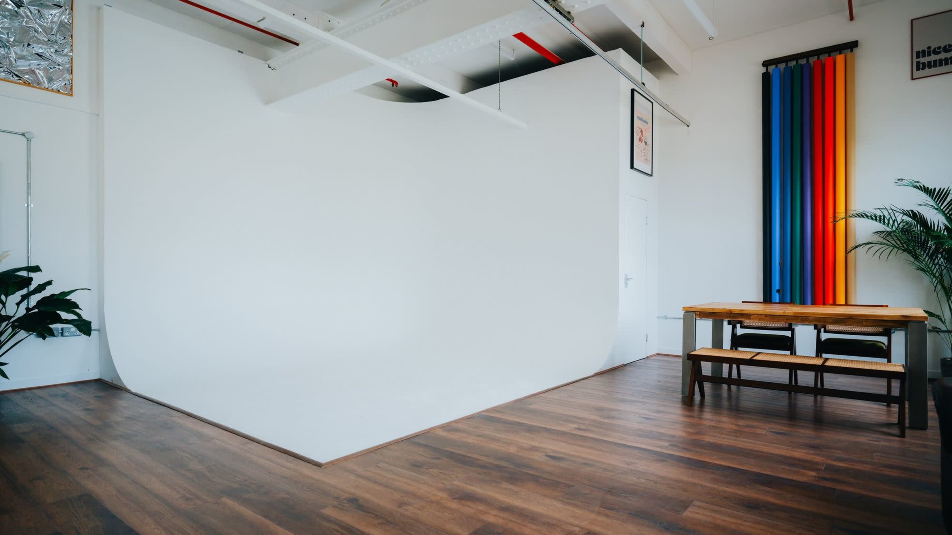 The image shows a bright studio space with a white backdrop, wooden flooring, a dining table with chairs, and colorful drapes on the wall.
