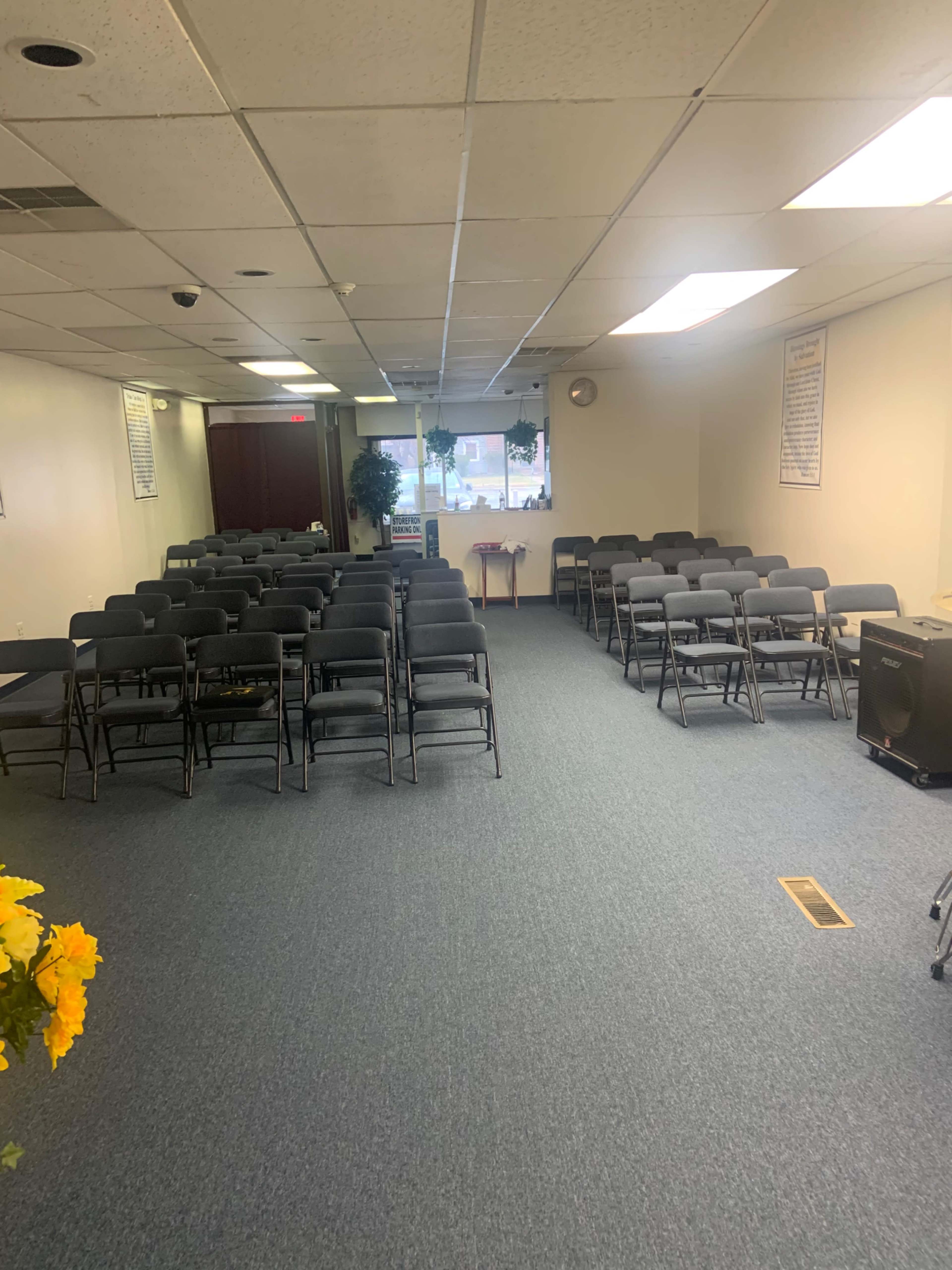 A rectangular room with rows of gray chairs set up for an event, a small table at the front, and windows allowing natural light.