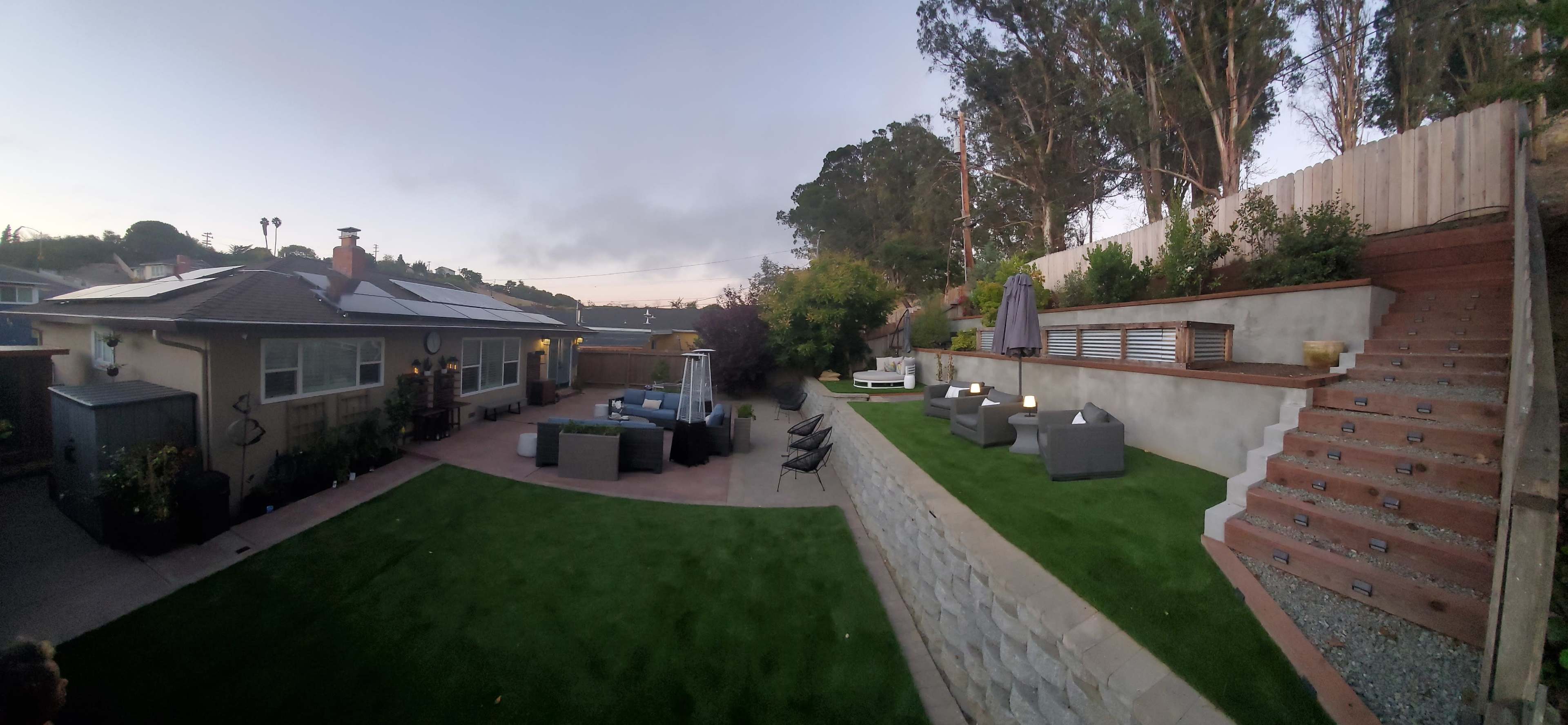 A landscaped backyard with seating areas, artificial grass, a staircase, and trees in the background.