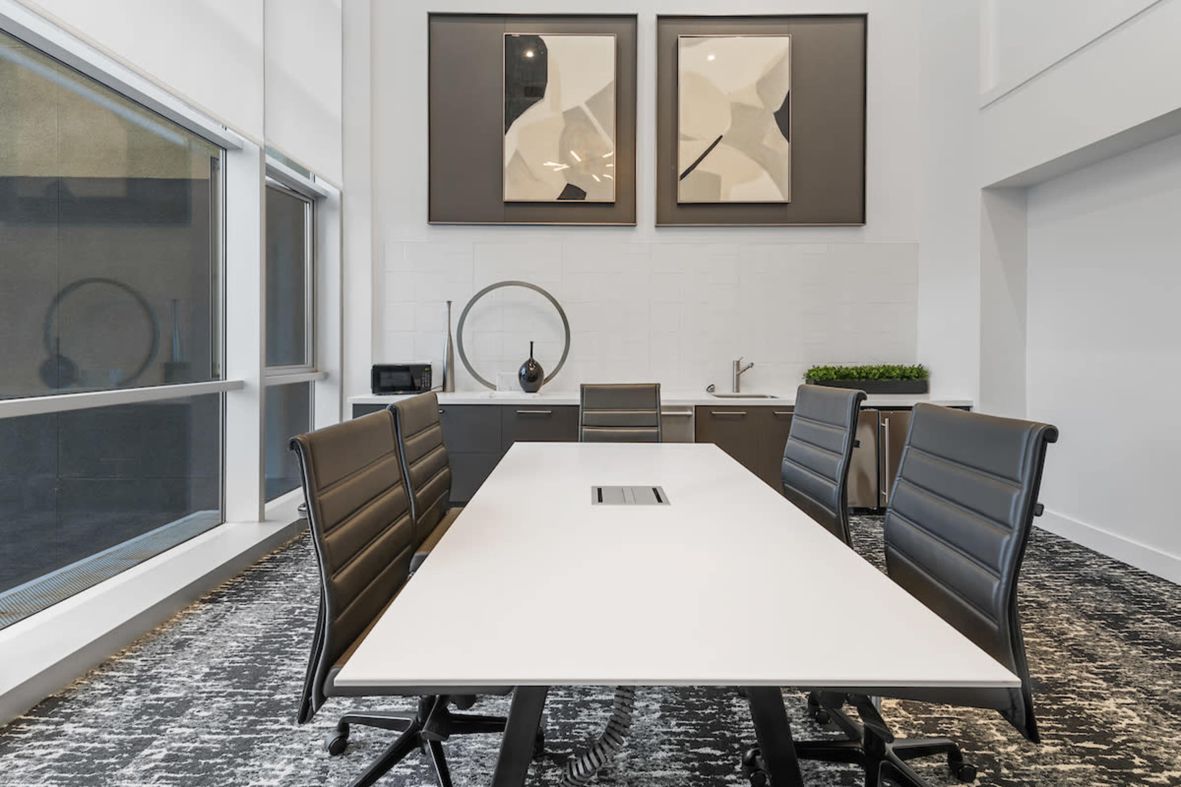 A modern conference room features a long white table surrounded by black leather chairs, with large windows and abstract artwork on the walls.