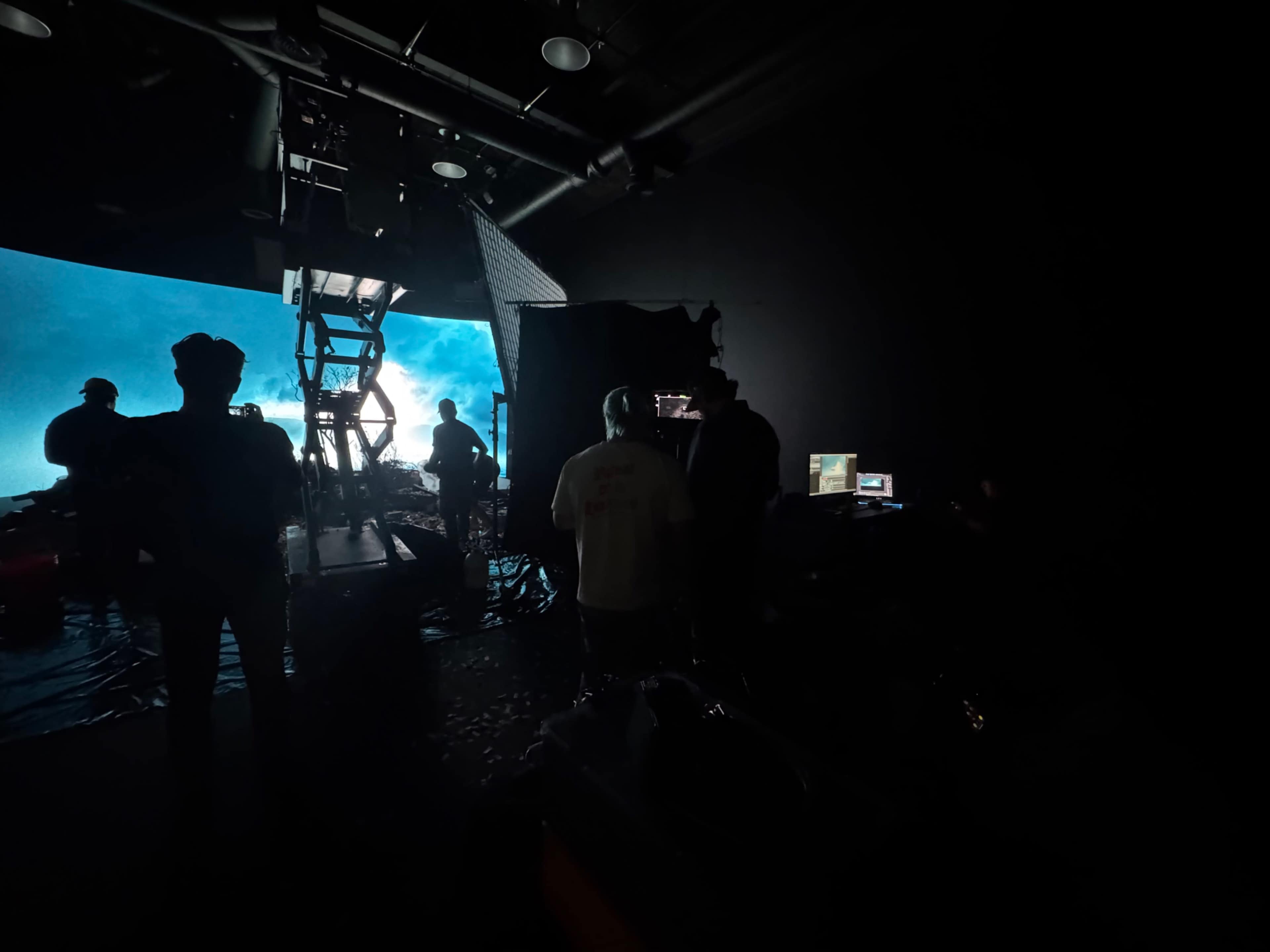 A group of people is working on a film set illuminated by a large screen displaying a blue and turquoise scene.