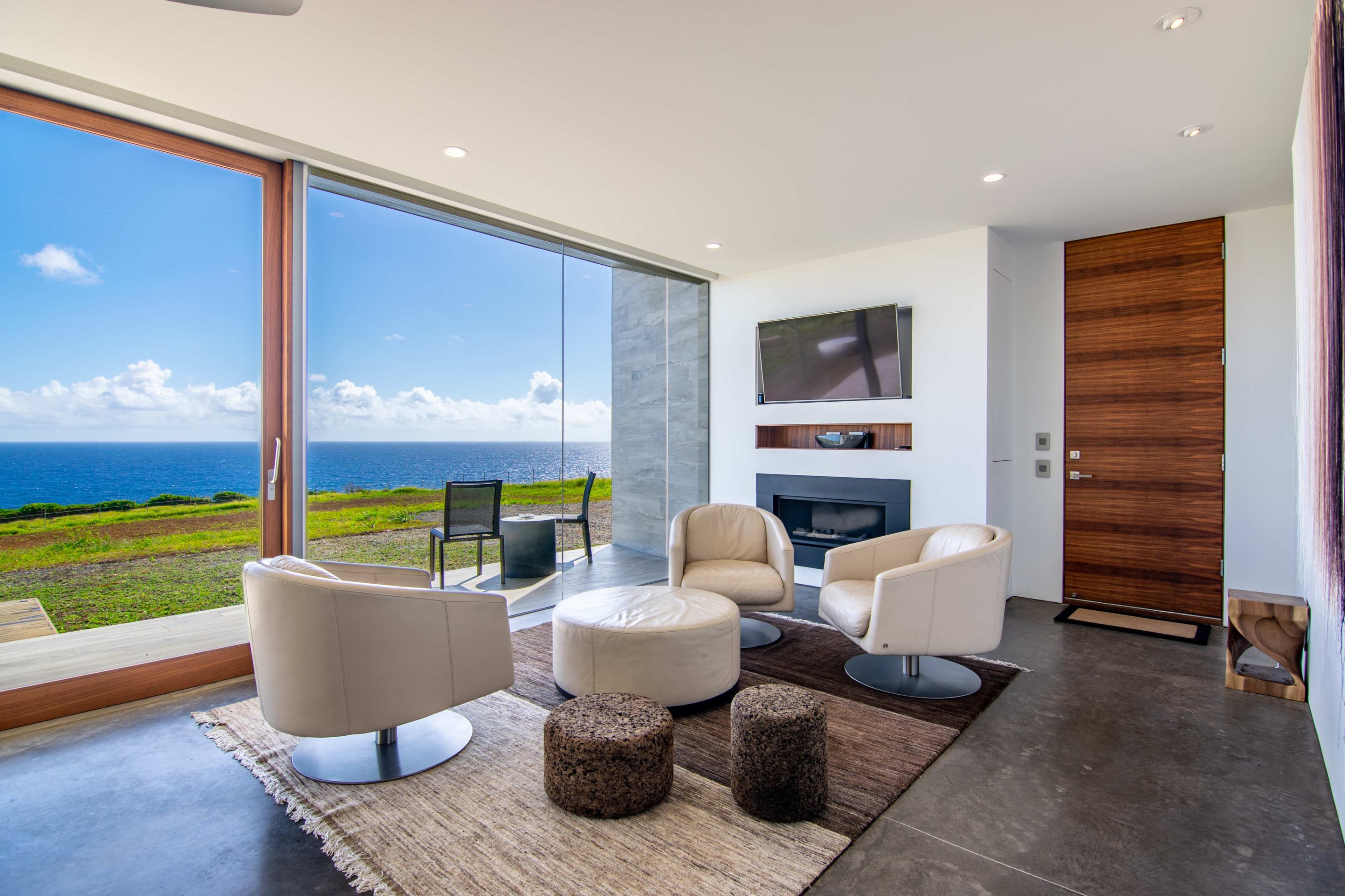 The scene depicts a modern living room with two white swivel chairs, a circular coffee table, a television mounted on the wall, and large windows offering a view of the ocean and sky.