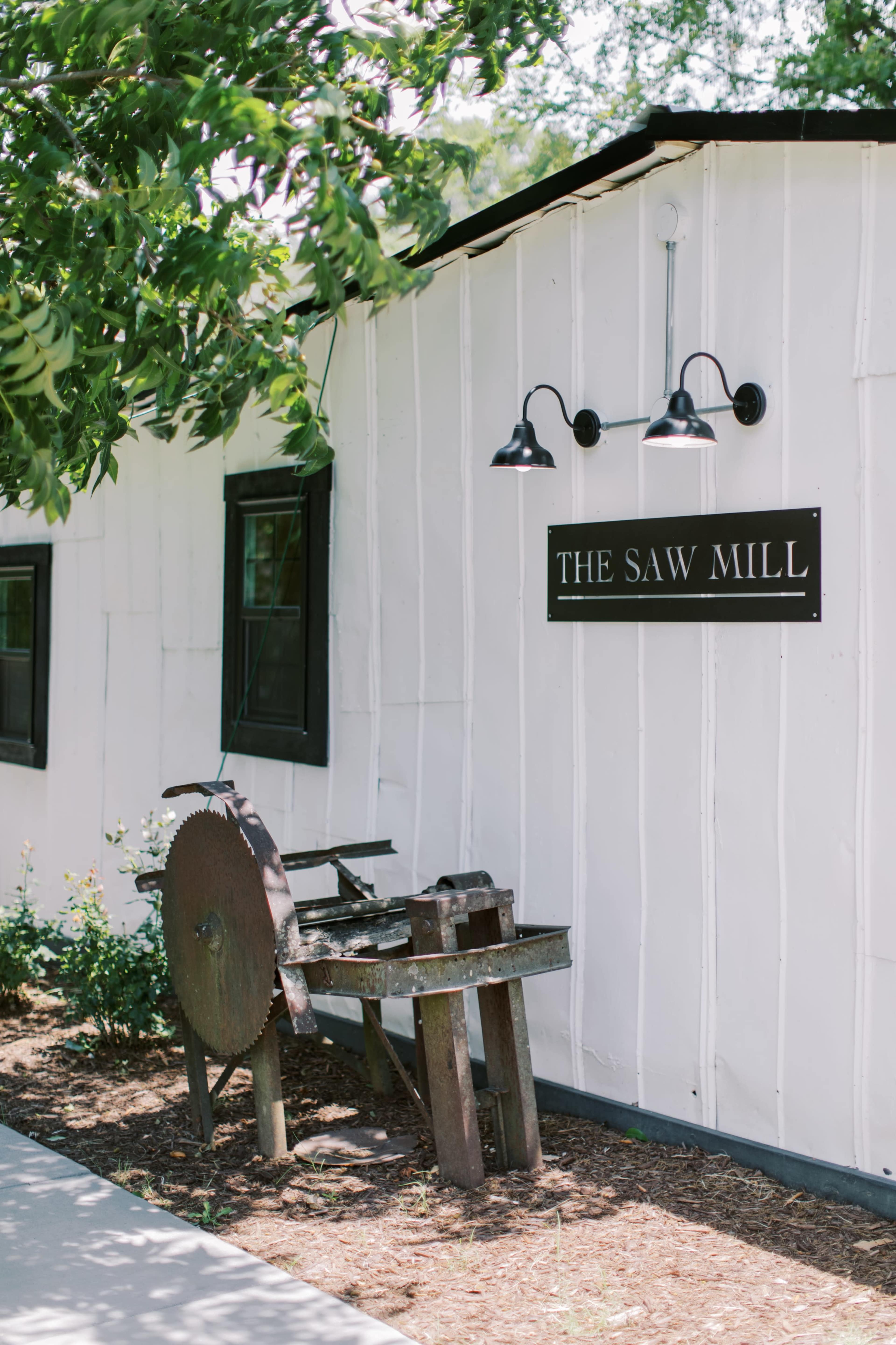 A vintage sawmill machine is positioned next to a white building with a sign that reads "THE SAW MILL."