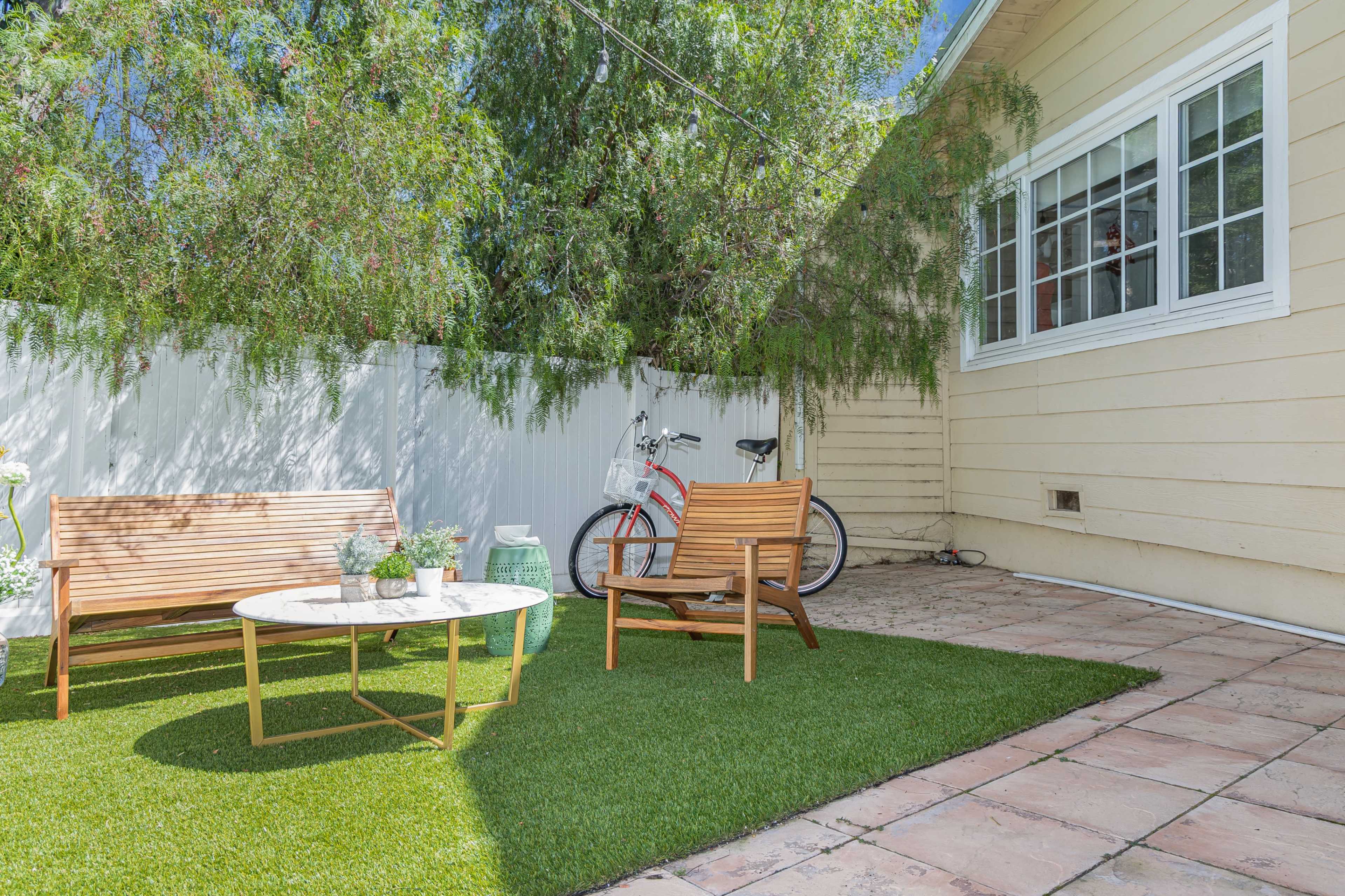 A small outdoor patio features two wooden benches, a round coffee table, and a bicycle leaning against a fence under a tree.