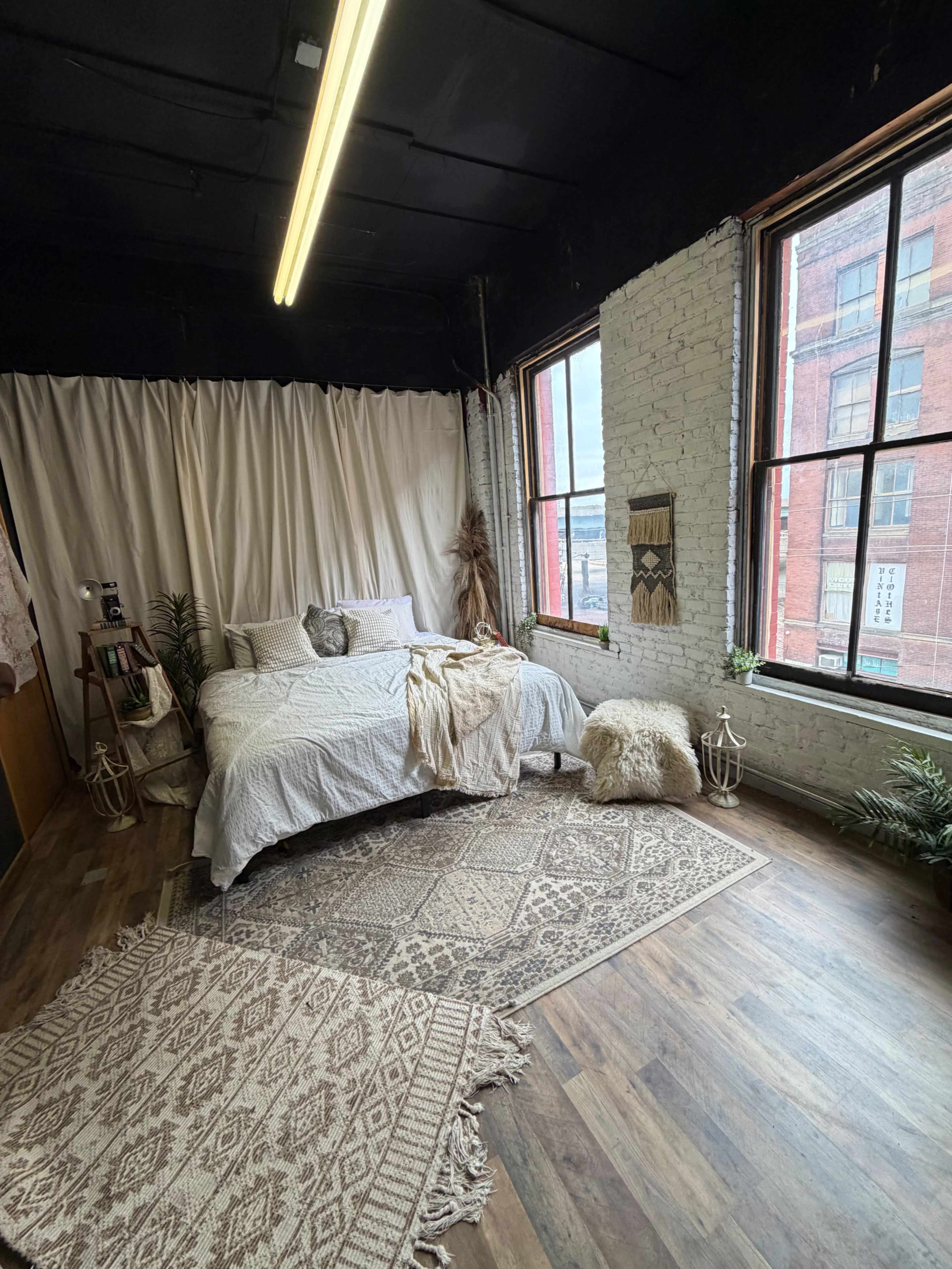 The image shows a cozy bedroom with a bed covered in light bedding, adorned with decorative throw blankets, surrounded by windows that let in natural light and featuring patterned rugs on a wooden floor.