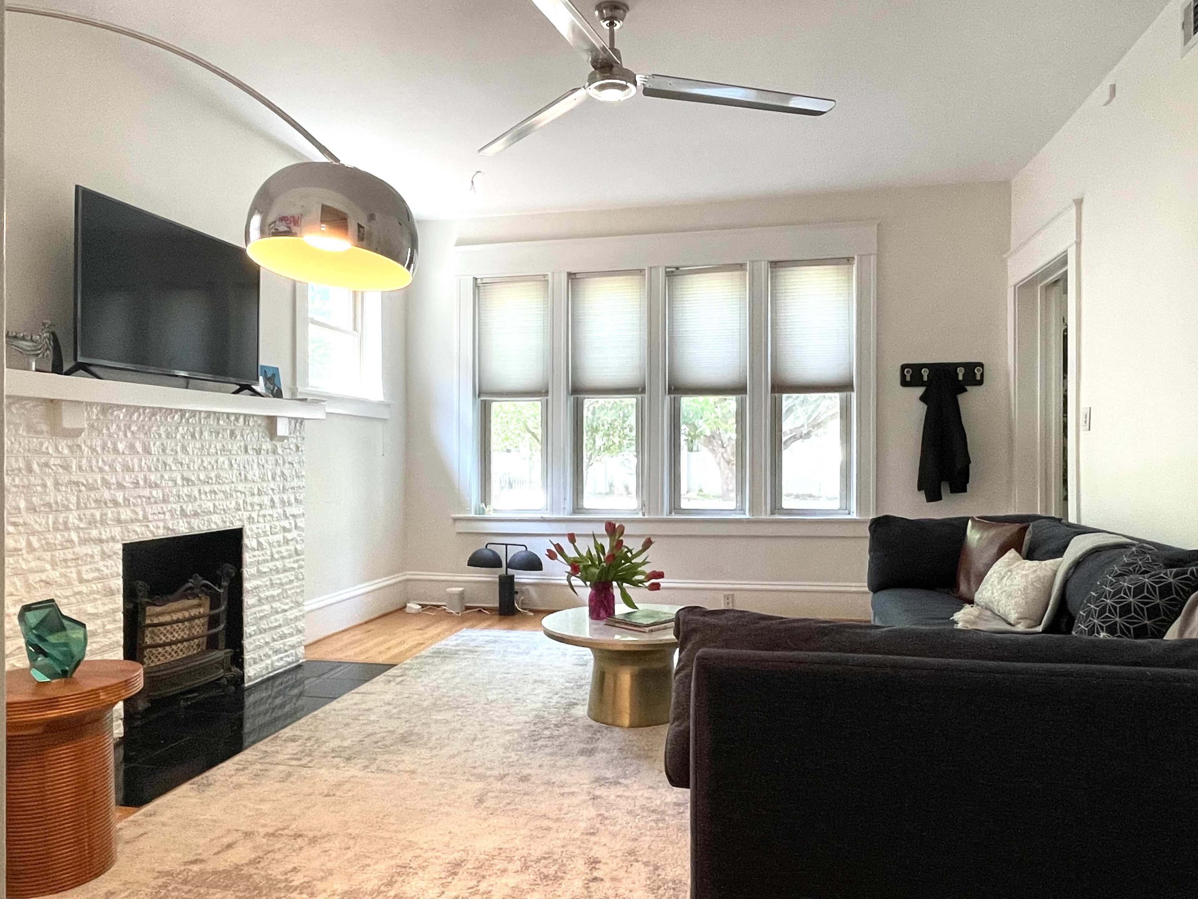 A cozy living room featuring a sectional sofa, a round coffee table with a floral arrangement, a modern lamp, and a flat-screen TV mounted above a white brick fireplace.